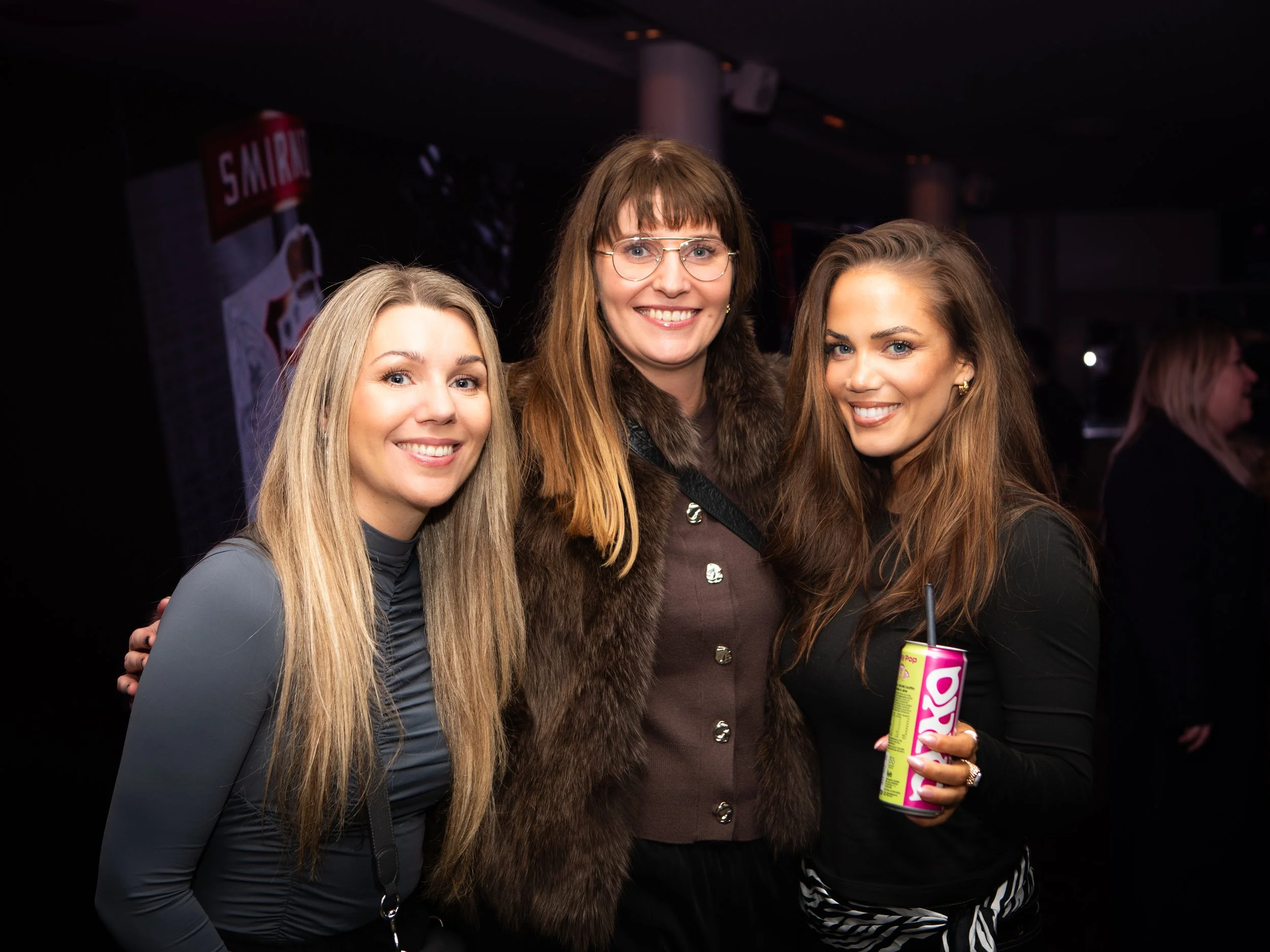Three women smiling at a social event, two of them with long blonde and brown hair, one with glasses and a fur-collared coat, the other holding a can of bubbly drink.