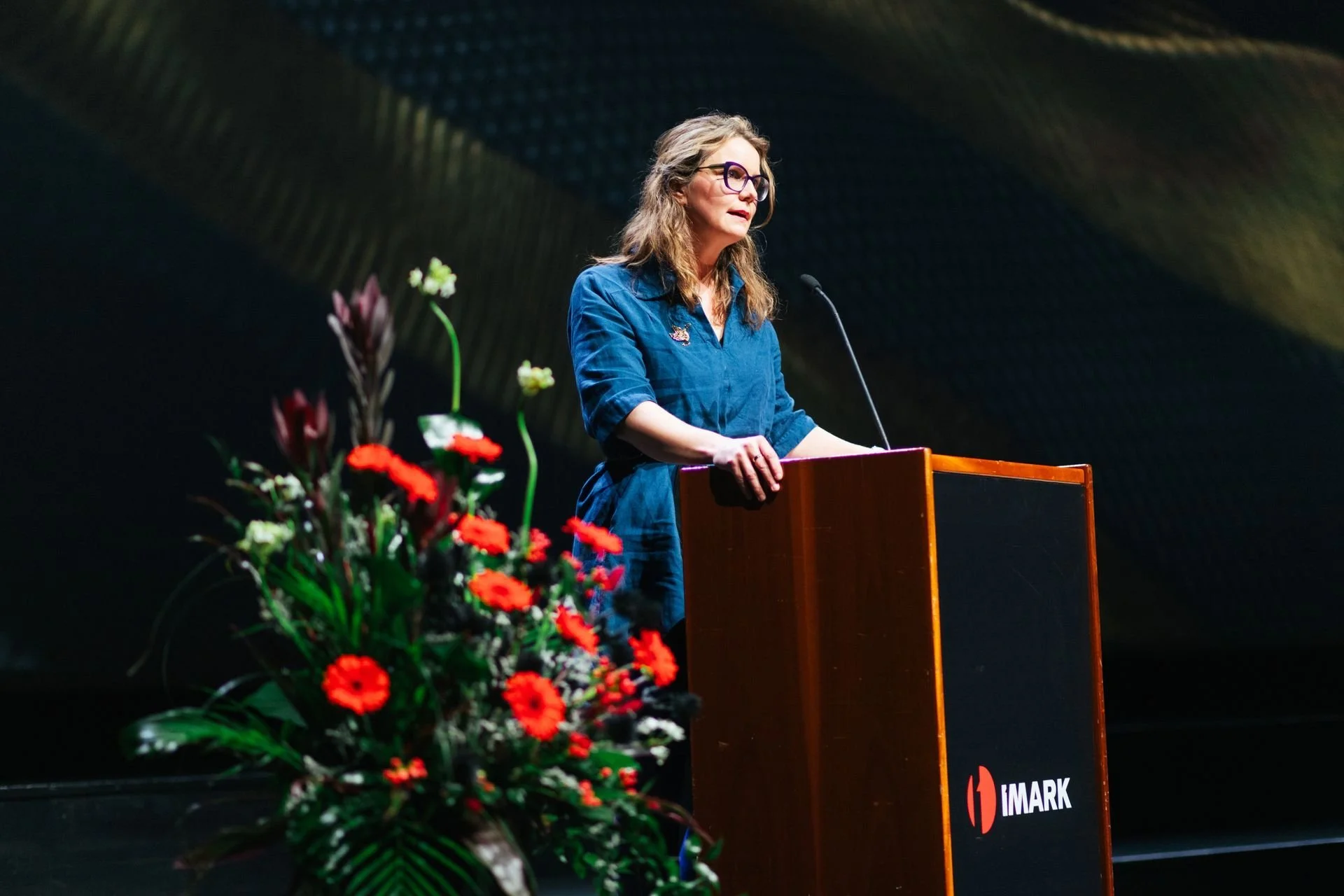 A woman with long gray hair and glasses stands at a podium, speaking into a microphone during a presentation or event. She is wearing a blue shirt and there is a large floral arrangement with red and white flowers in the foreground. The background is