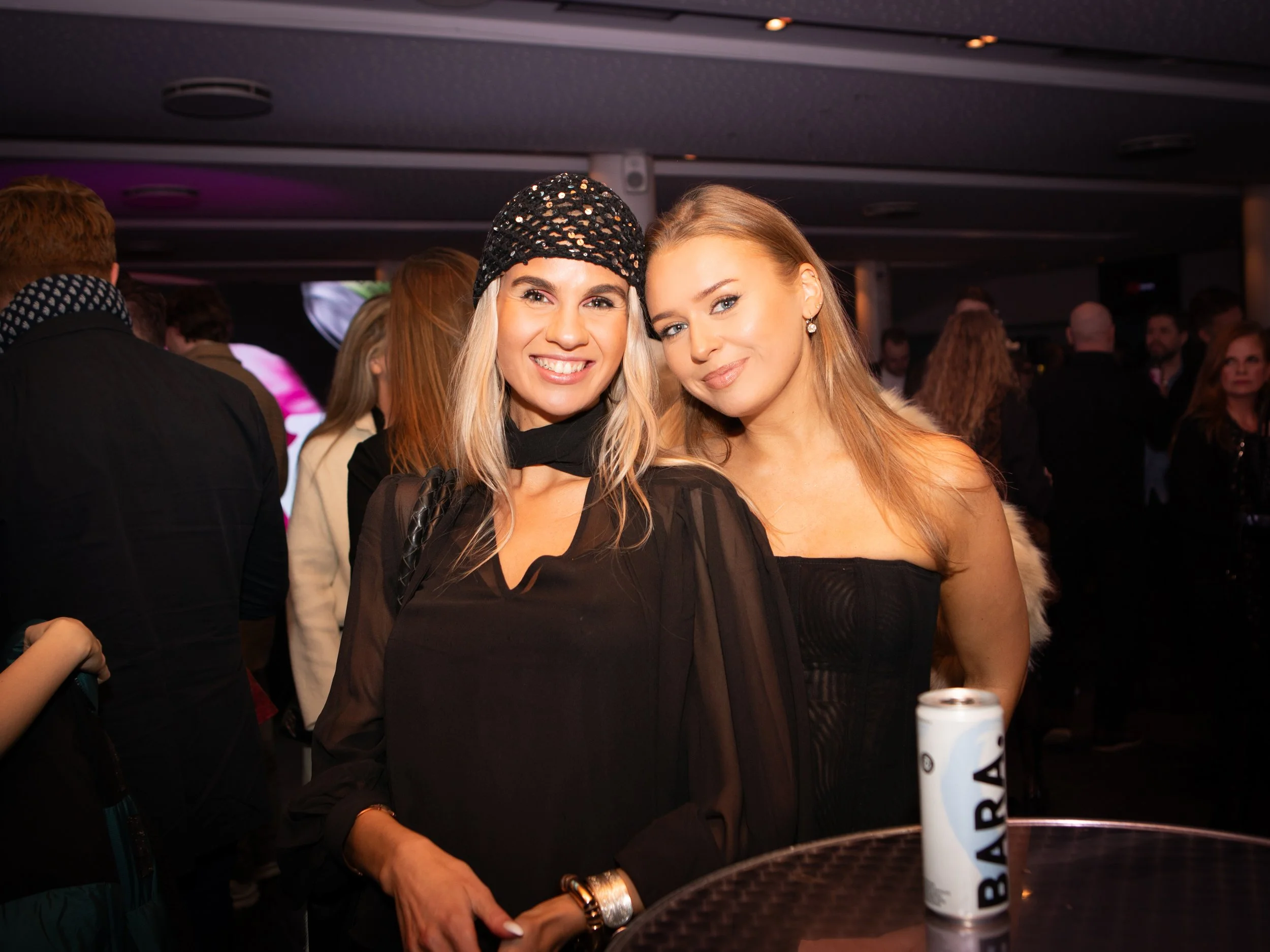 Two women smiling at the camera in a crowded indoor setting. The woman on the left is wearing a black sequined hat and a sheer black top with a black scarf. The woman on the right has long blonde hair, is wearing earrings, and a black dress. There ar