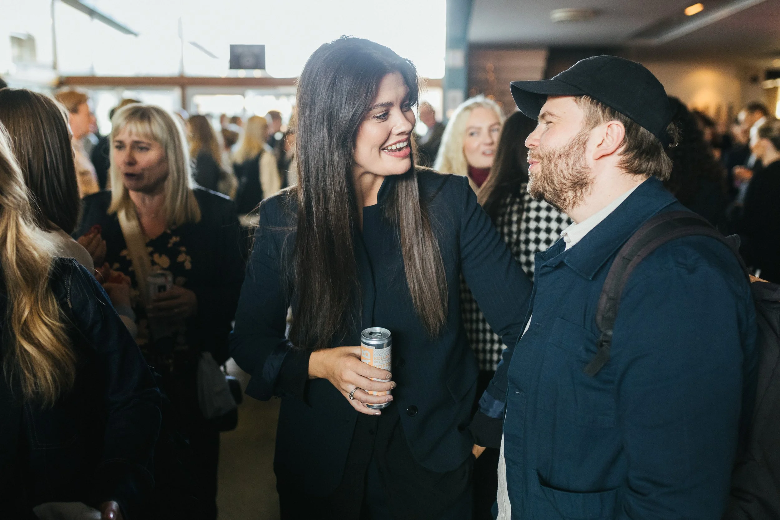 A woman with long dark hair wearing a black blazer is smiling and talking to a man in a dark blue jacket and black cap at a social gathering.