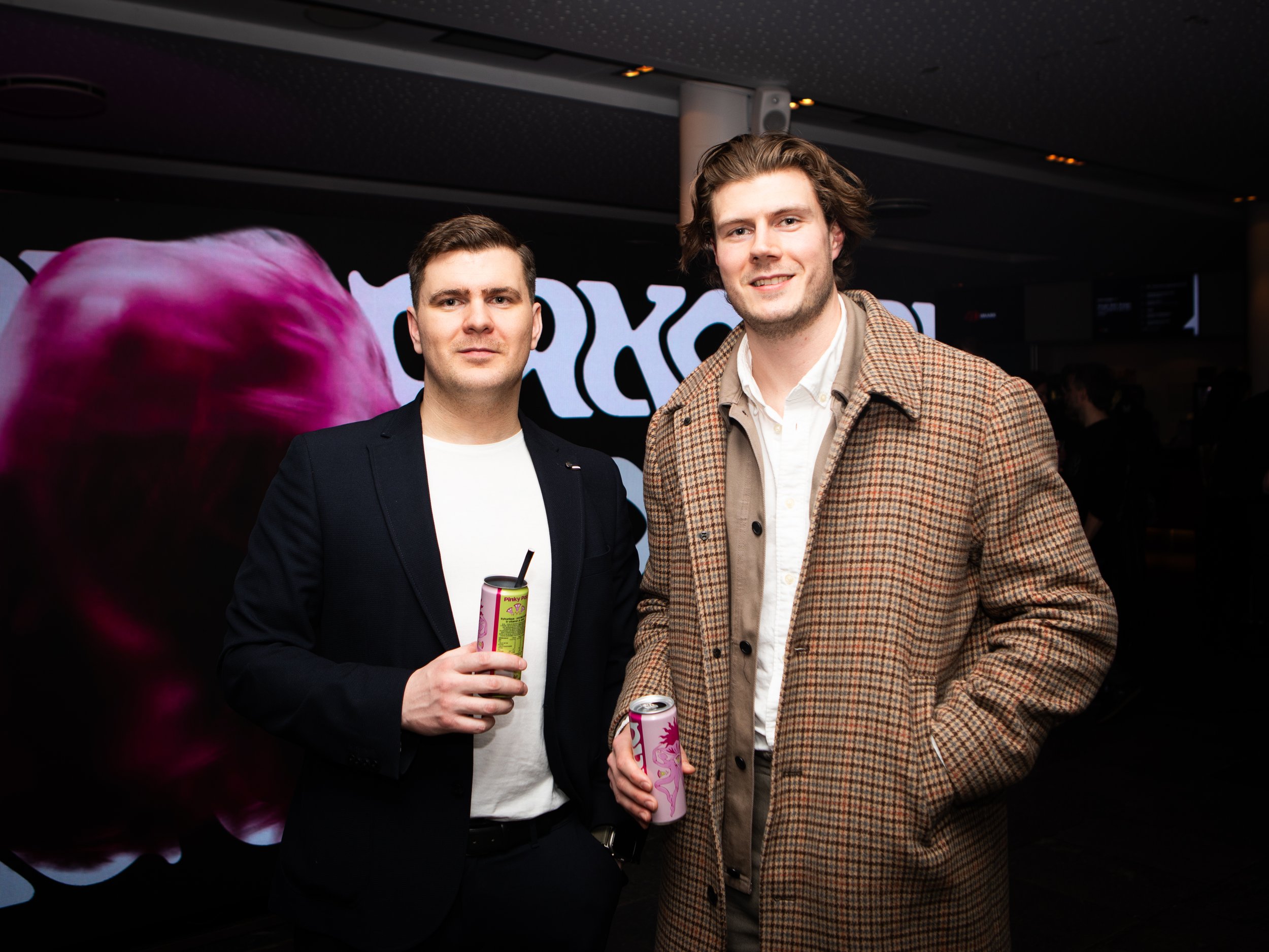 Two young men are standing together at an indoor event, holding drinks in their hands. One is wearing a dark blazer over a white shirt, and the other is wearing a checked brown blazer over a white shirt. They are smiling at the camera with a large, i