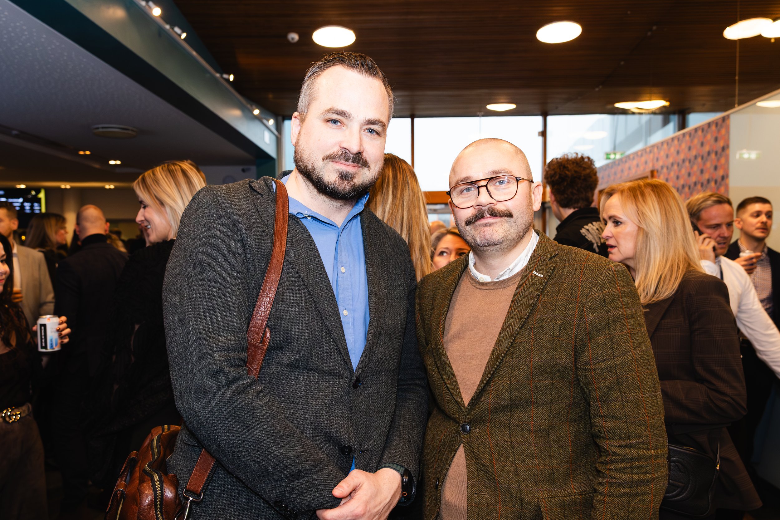 Two men standing closely together at a social event, smiling at the camera, with a crowded indoor setting in the background.