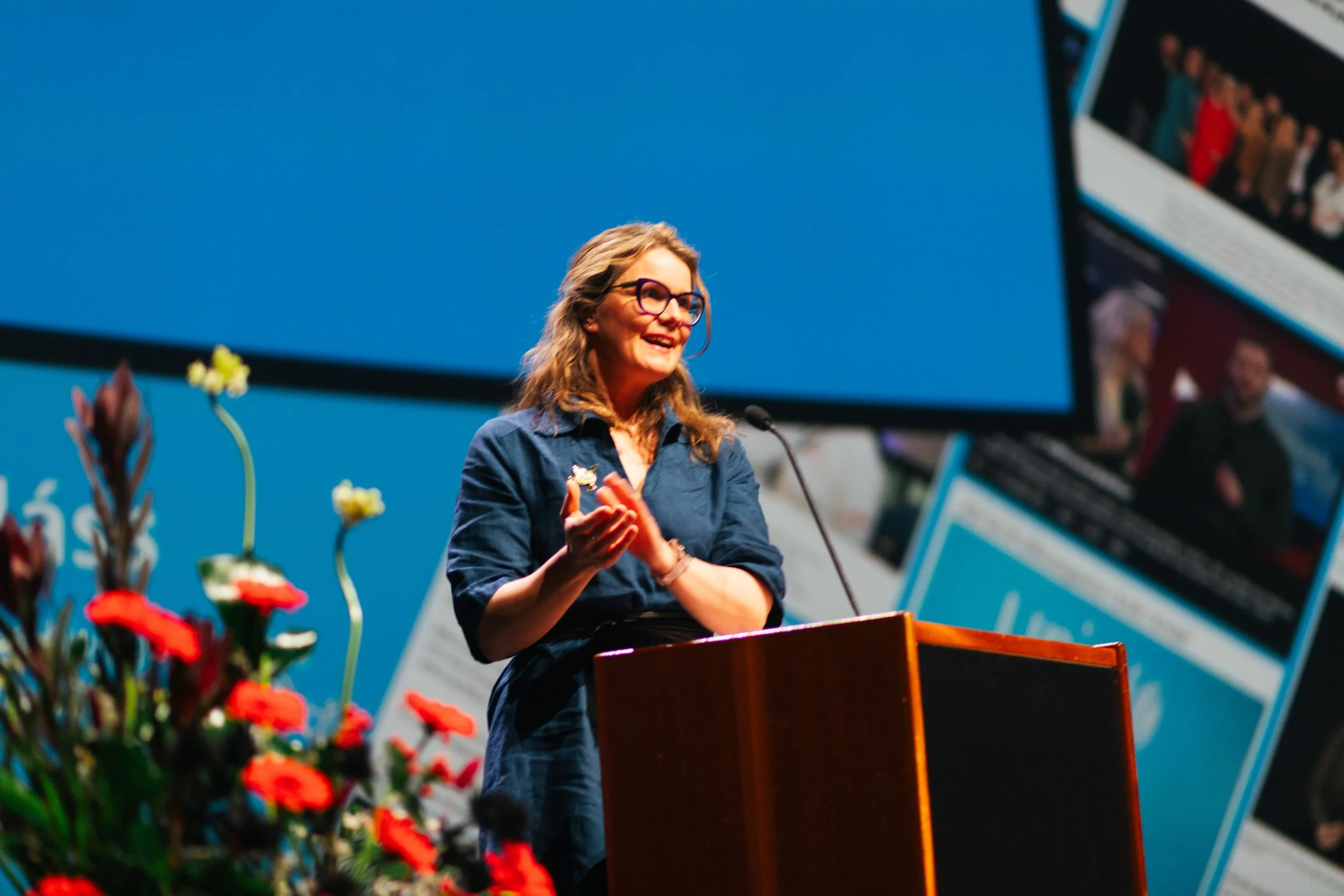 A woman with glasses and wavy hair, wearing a blue shirt, is standing behind a podium and speaking at an event. There are colorful flowers in the foreground, and a large screen and a busy background with people are visible behind her.
