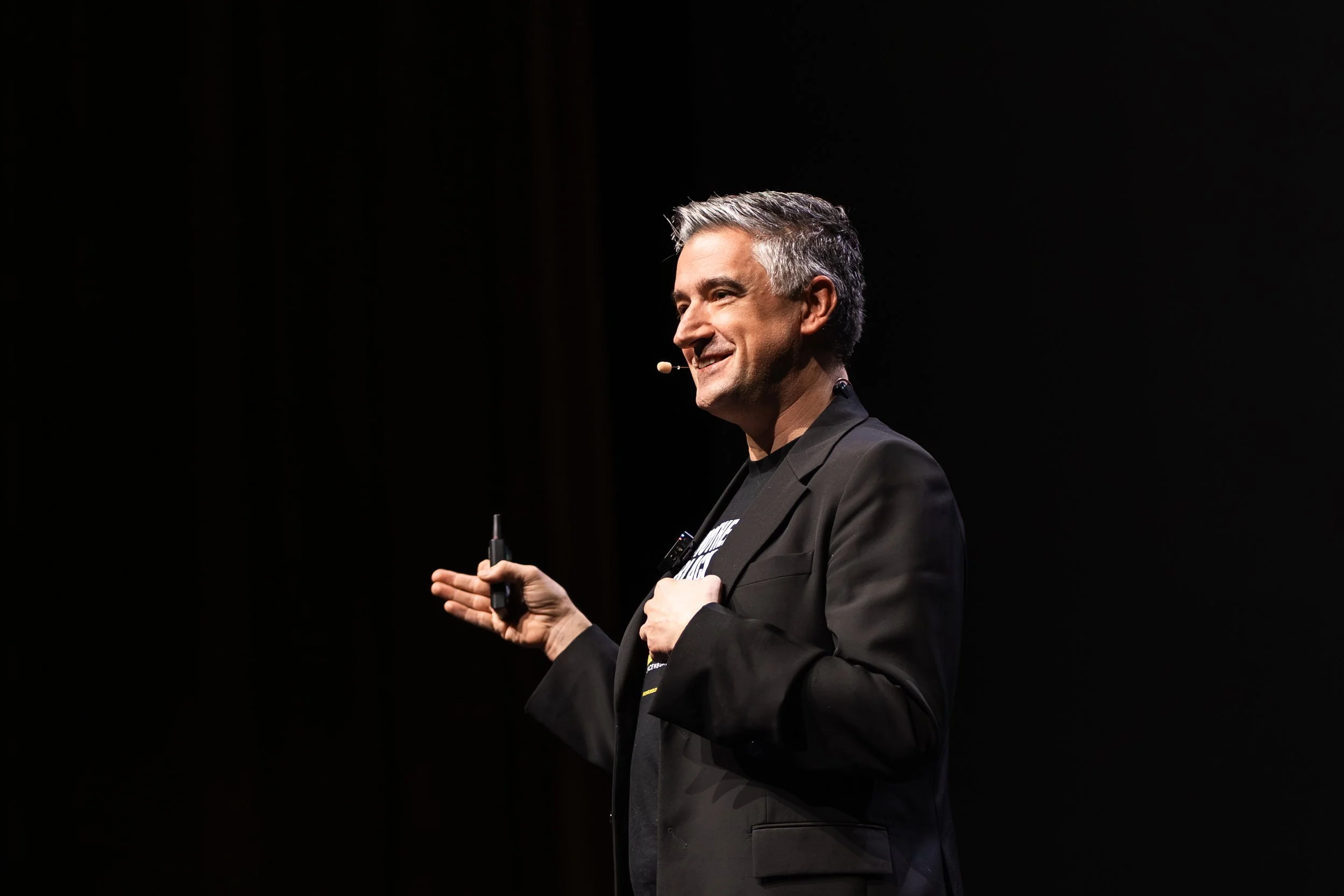 A man with gray hair, wearing a black suit and a headset microphone, smiling during a presentation or speech on a dark stage.