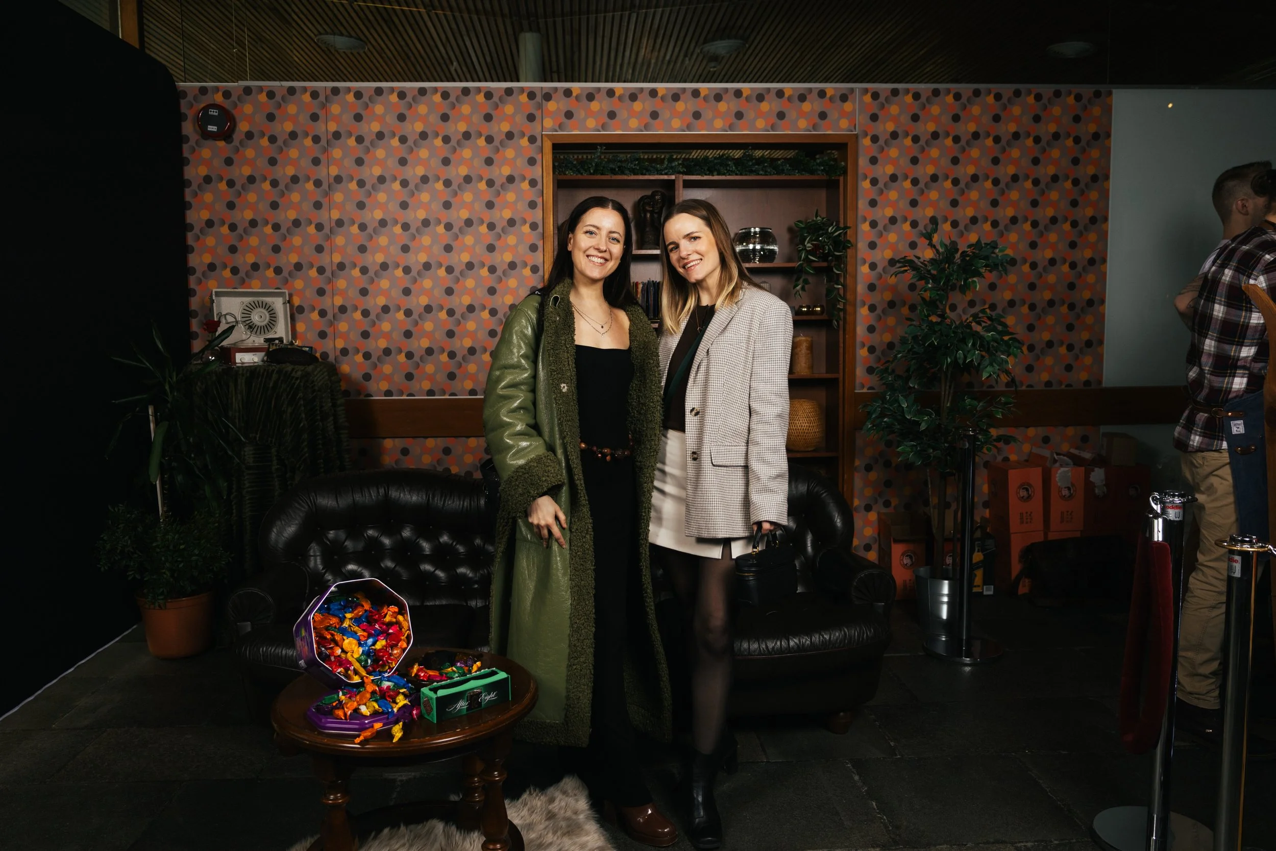 Two women smiling and standing together in a room with a colorful dotted wallpaper. There is a table with wrapped candies in front of them and a black leather sofa behind them. One woman is wearing a green jacket, and the other is wearing a plaid bla