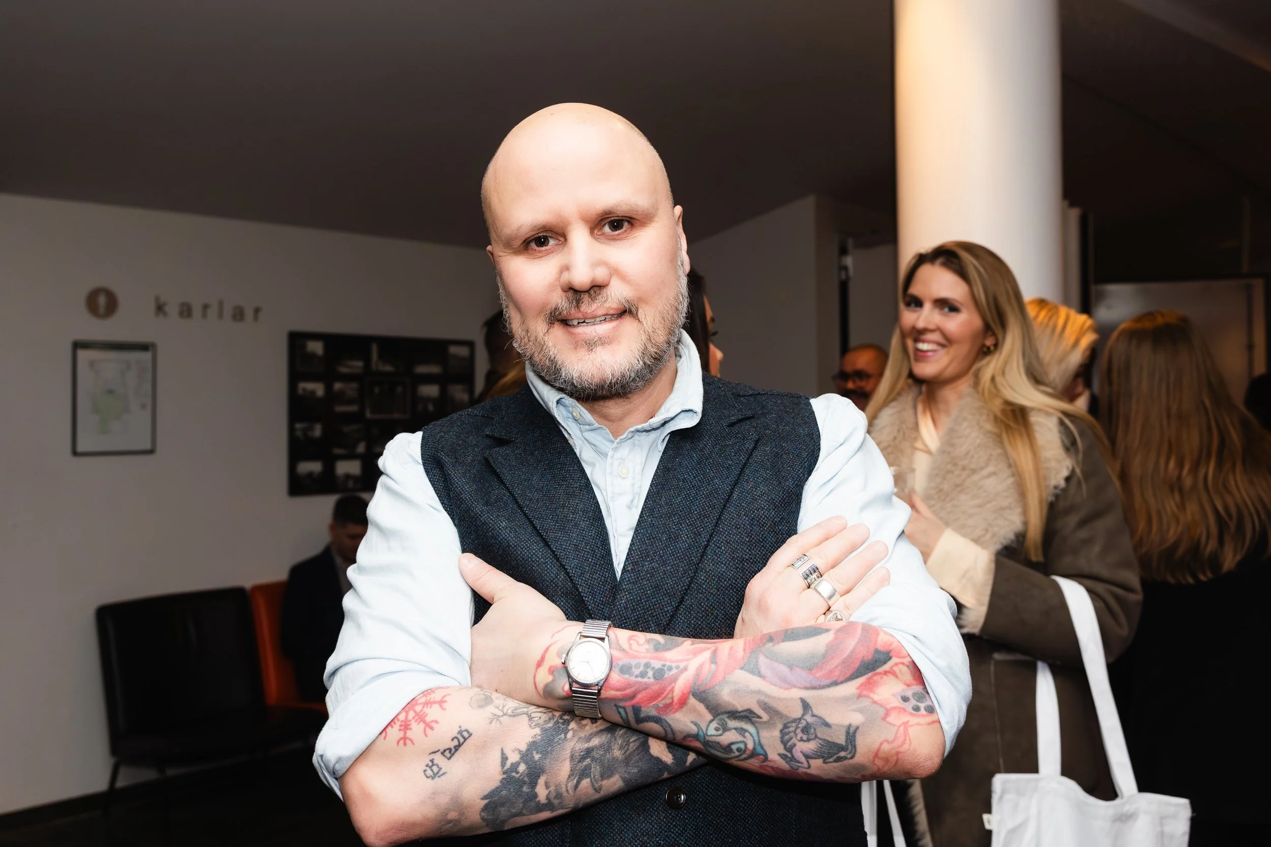 A bald man with a beard, dressed in a light blue shirt and dark vest, crossing his arms with tattoos visible on his forearms, standing in an indoor setting at an event or gathering. There are other people and a white column in the background.