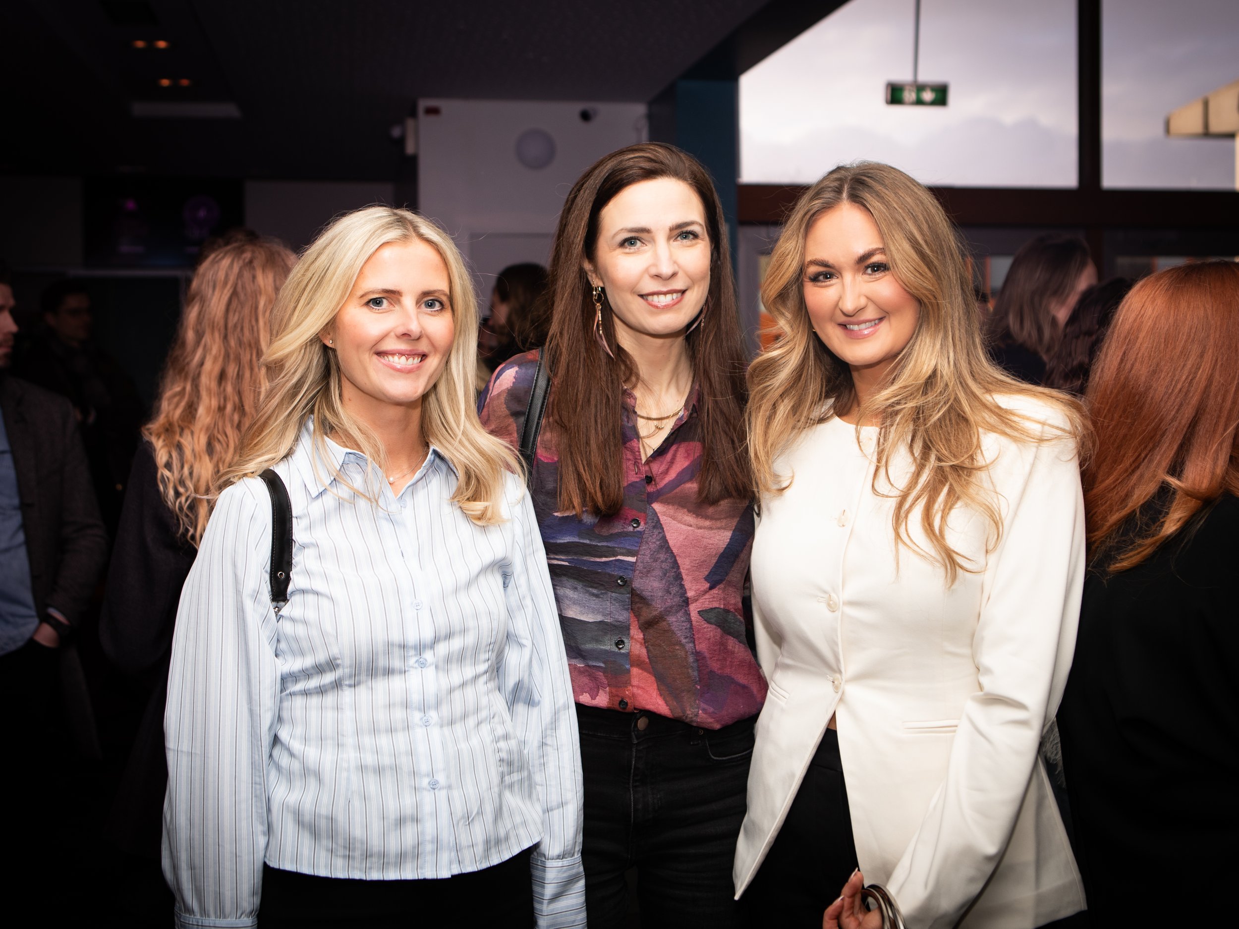 Three women smiling at a social event, standing close together in an indoor setting with other people in the background.