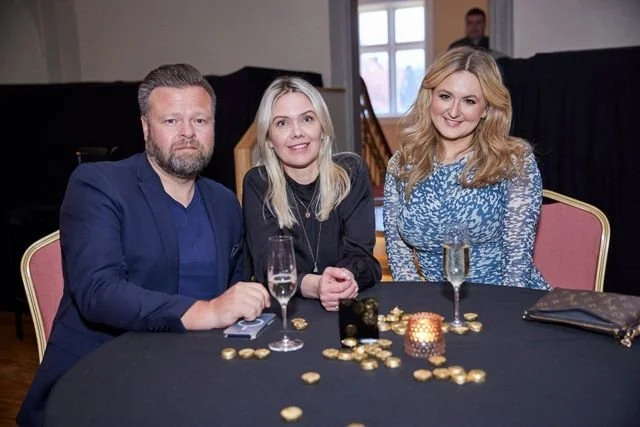 Three people sitting at a black table with gold coins and a lit candle, in a room with large windows and a man standing in the background.