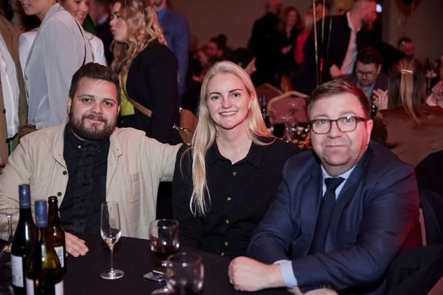 Three people sitting at a table in an event or party, smiling at the camera. On the table are several bottles and glasses of drinks.