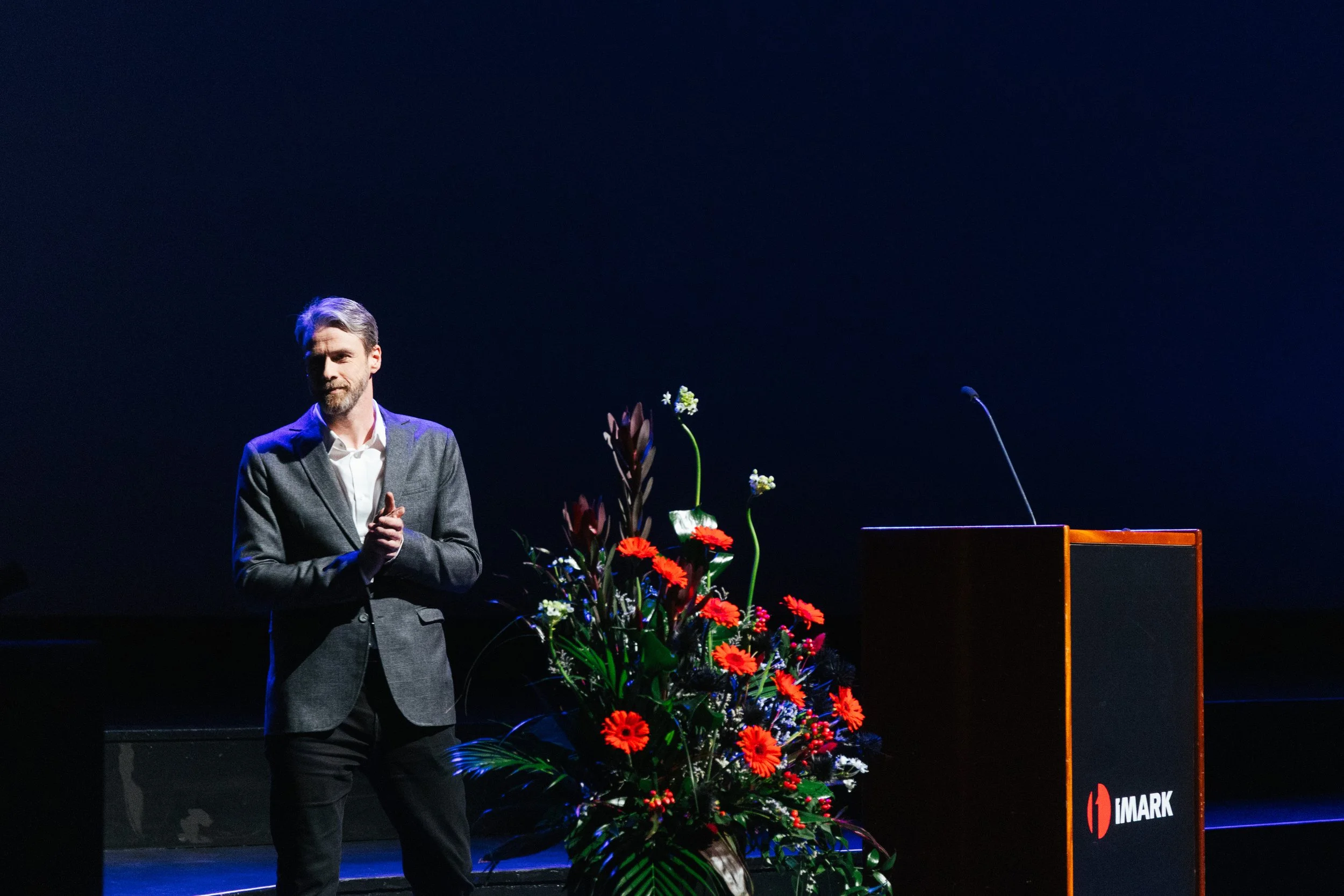 A man in a gray suit is standing on stage with a dark background, next to a large bouquet of red flowers and a lectern with the IM4ARK logo.