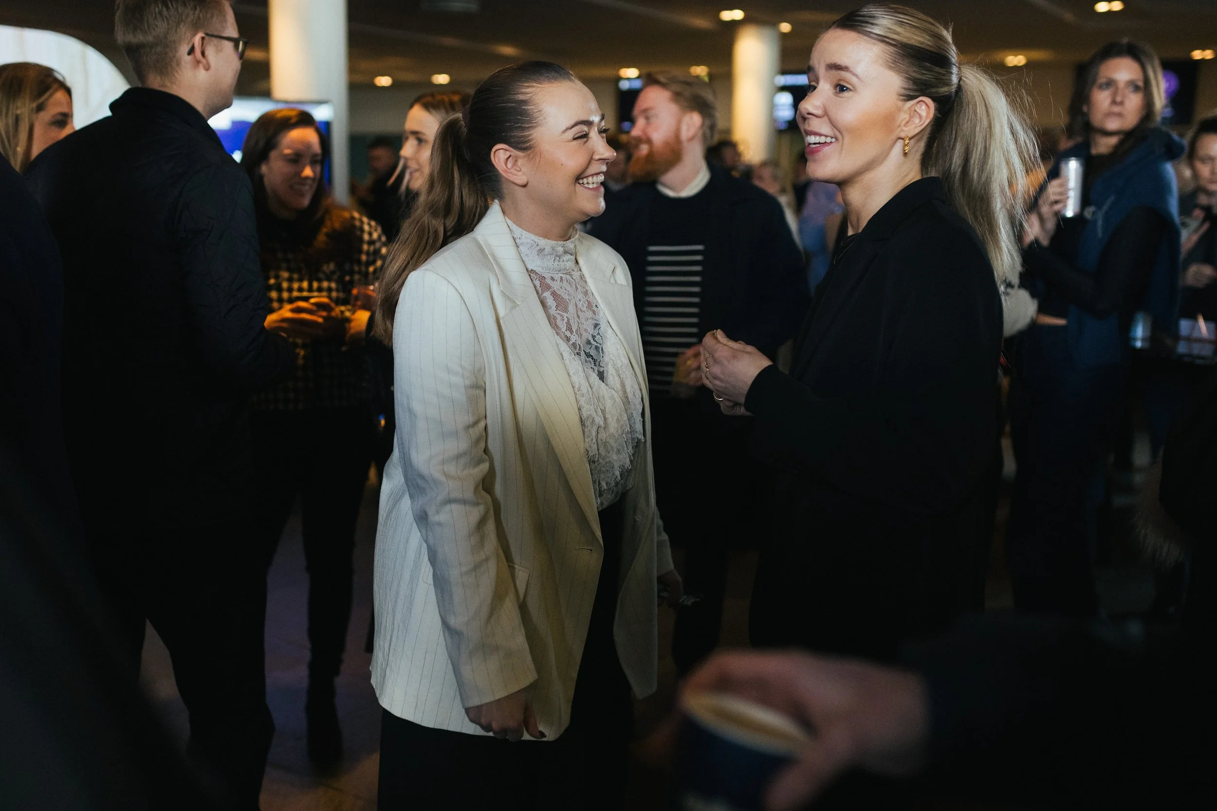 Two women smiling and talking to each other at a social event, with other people in the background.