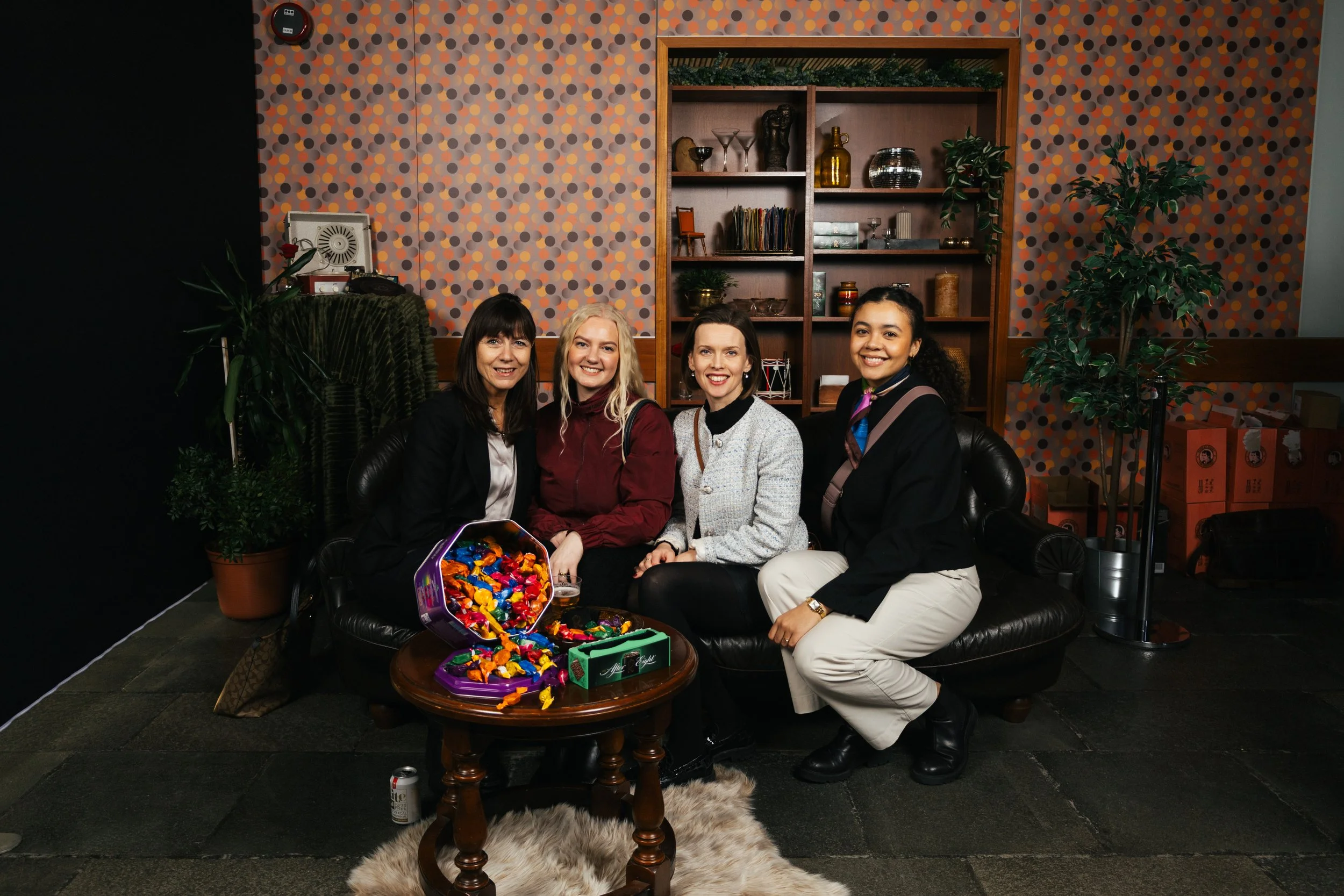 Four women sitting on a black leather couch, smiling at the camera, with a round wooden table in front of them filled with assorted chocolates and snacks. The background includes patterned wallpaper, a wooden shelf with decorative items, and plants.