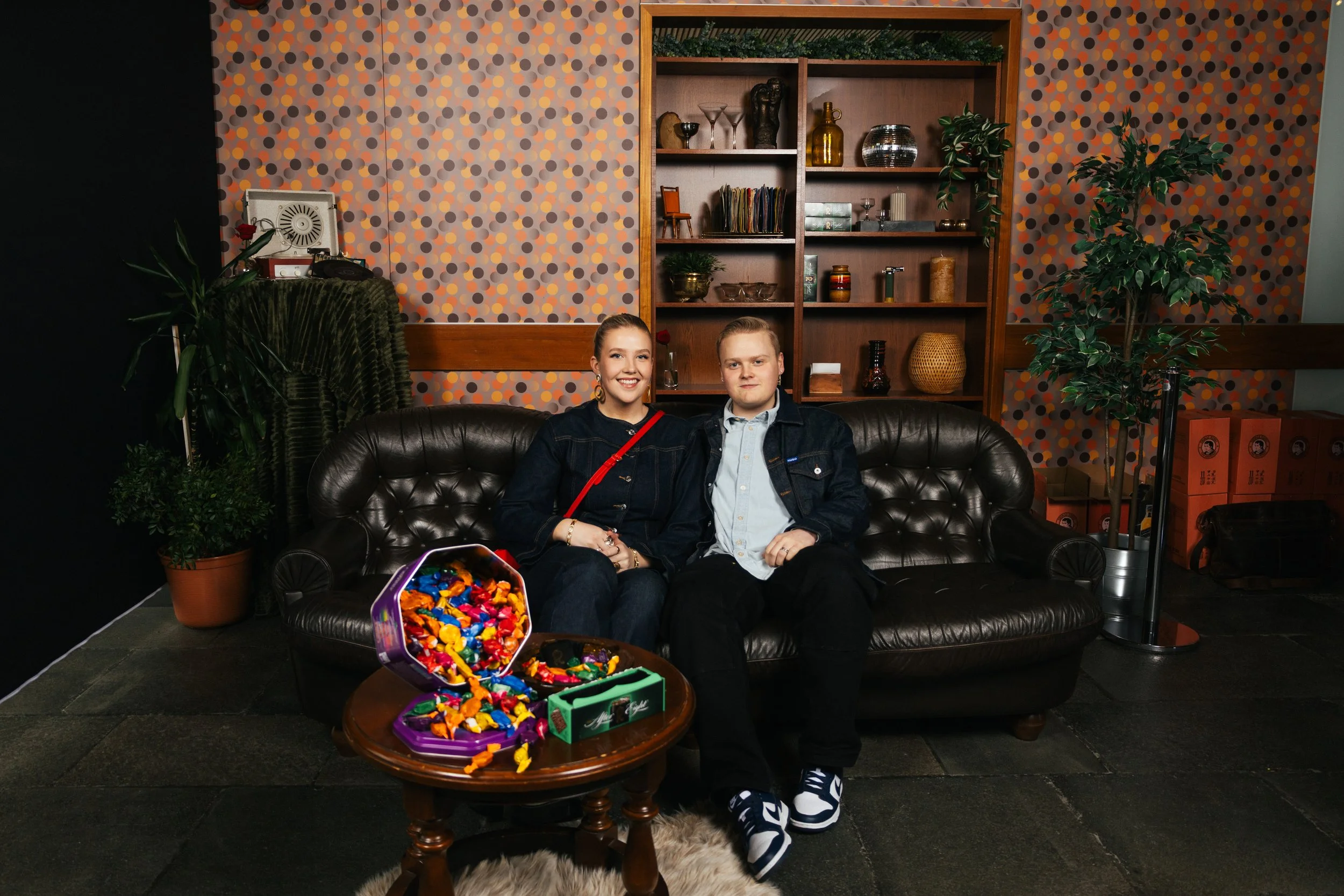 Two young people, a girl and a boy, sitting on a black leather couch in a cozy indoor setting, smiling at the camera. In front of them is a small round wooden table with a purple toy box open, filled with colorful candies. The background features a p