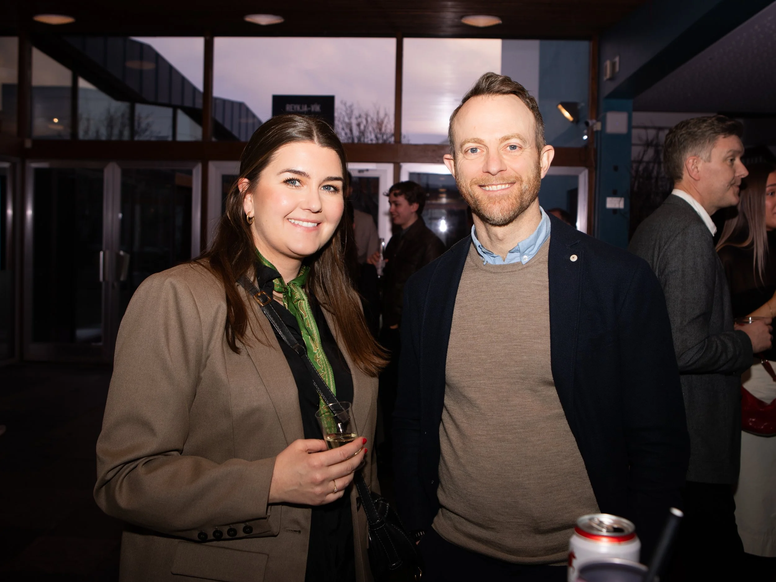A woman with long dark hair and blue eyes holding a glass of champagne, standing next to a man with short light brown hair and a beard, both dressed in semi-formal attire at an indoor social event.