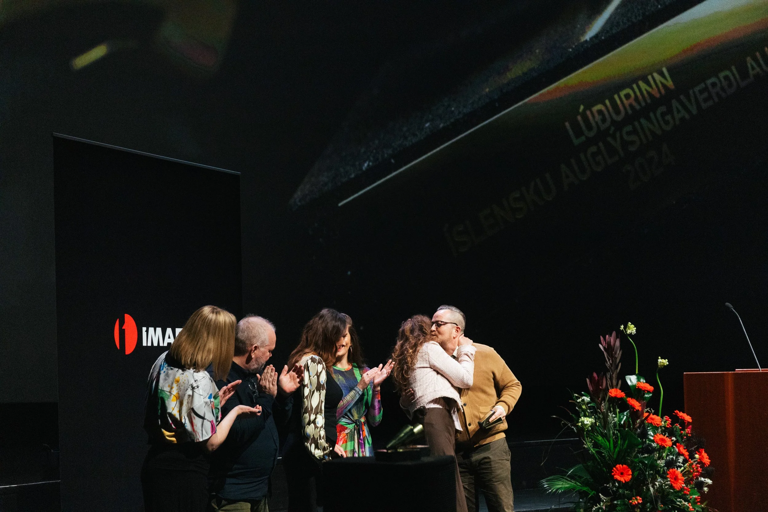 A group of people standing on stage at an event, with a black backdrop displaying the IMAX logo, some flowers on the right side, and text about an exhibition in Lugano, Switzerland, from August 20, 2023.
