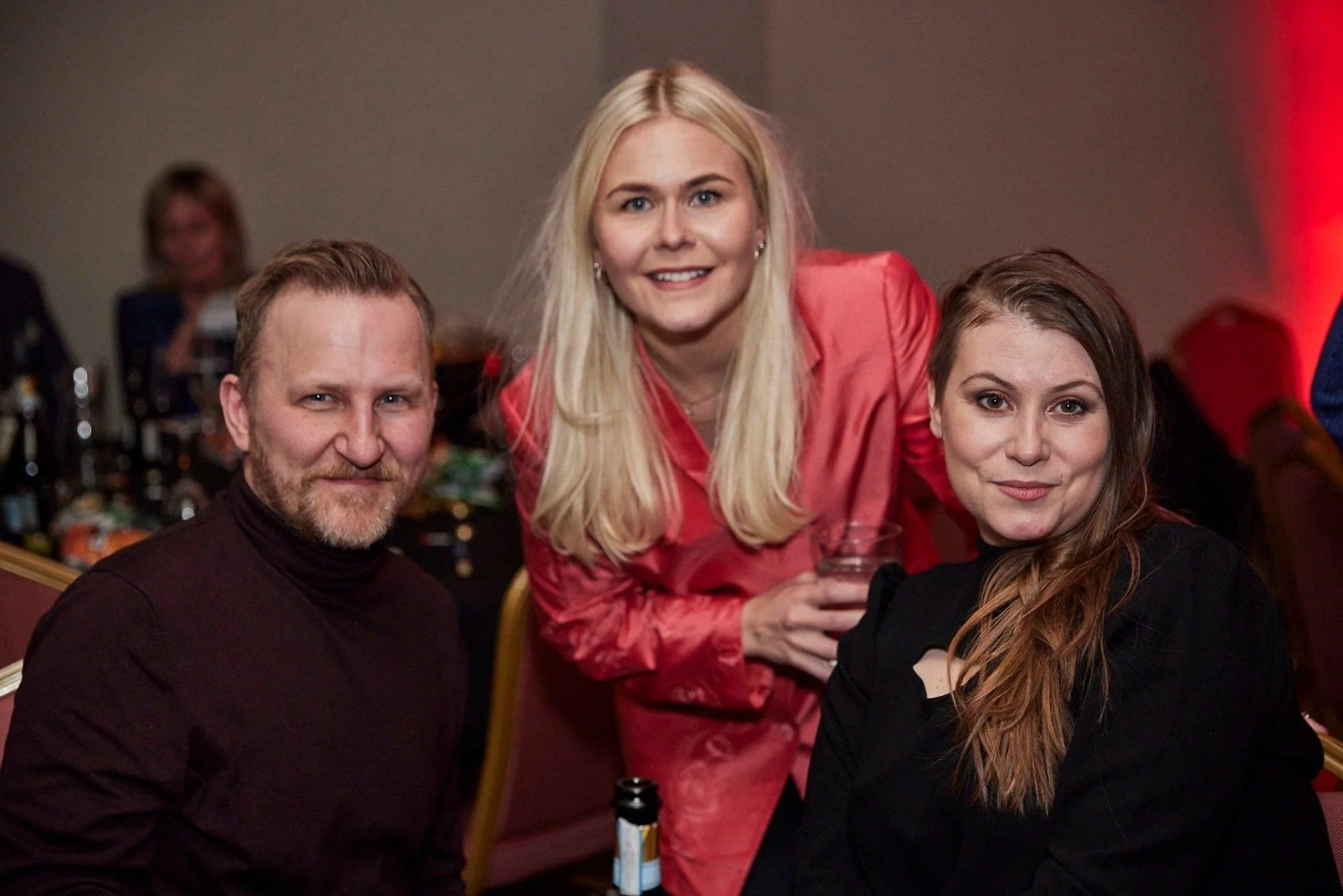 Three people at a social gathering, with two women and one man in the foreground. The woman in the middle, wearing a red satin jacket, is smiling and holding a glass, while the man and the woman next to her are also smiling, seated at a table with dr