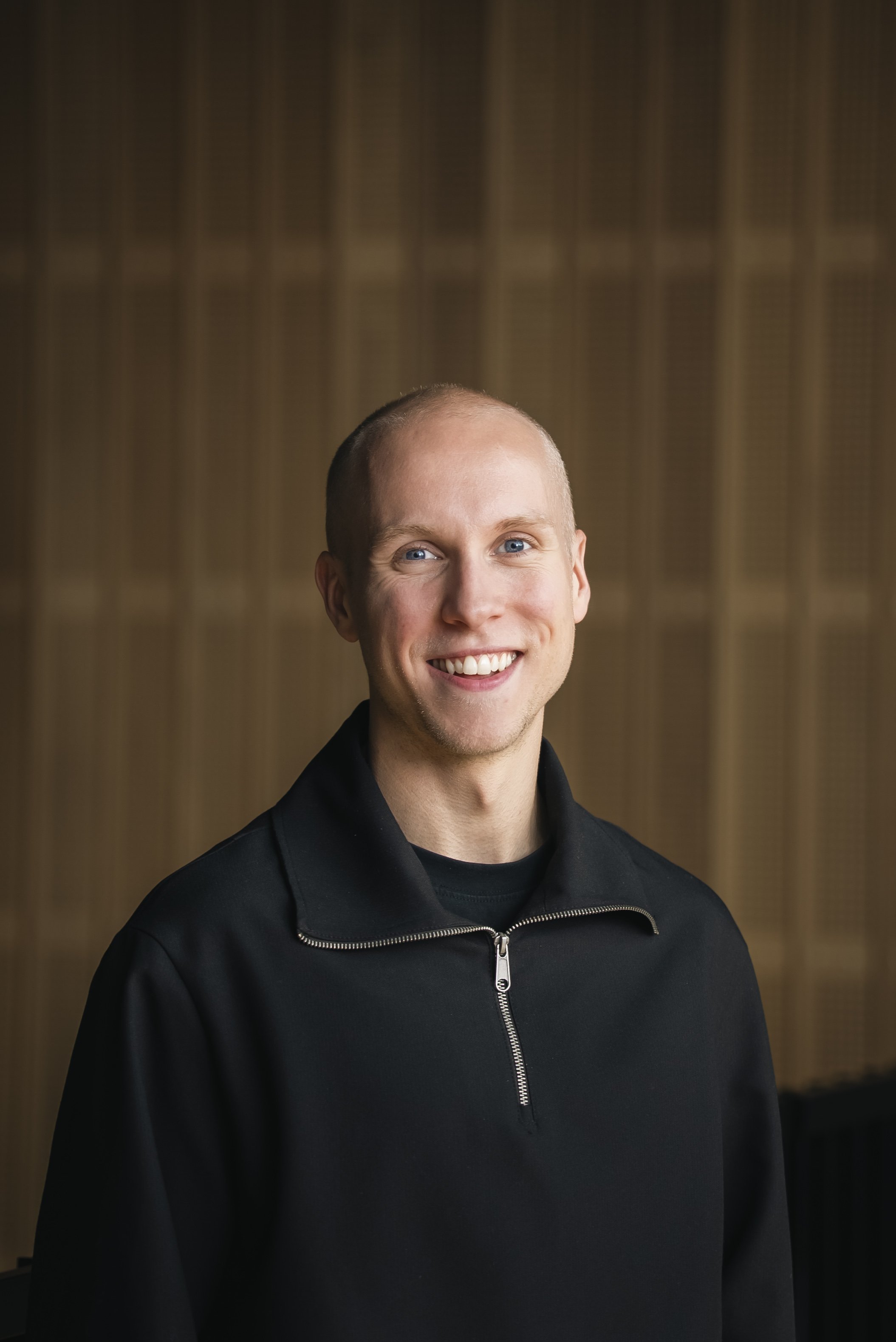 A young man with a shaved head and blue eyes smiling, wearing a black zip-up jacket, standing in front of a wooden panel background.