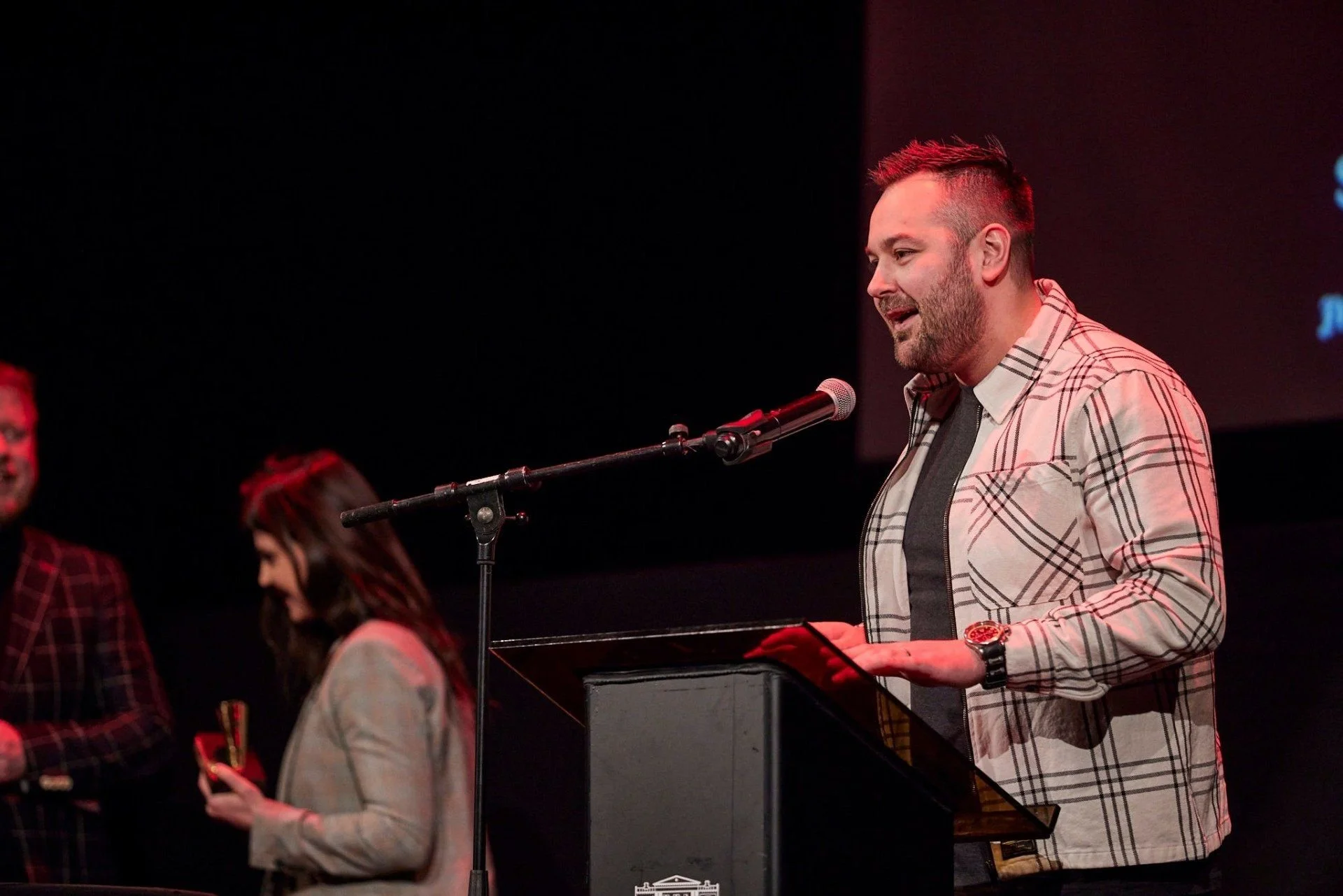 A man with short hair and a beard, wearing a plaid jacket and watching a screen, is speaking into a microphone and standing at a podium. Two women are seated nearby, one with long hair looking at her phone, and another with short hair observing. The 