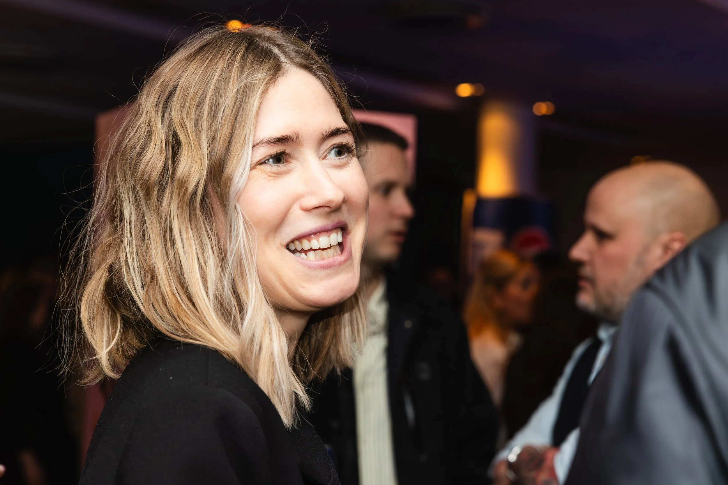 A woman with blonde hair smiling at an event with a dark background, people are seen in the background, including a man with a shaved head.