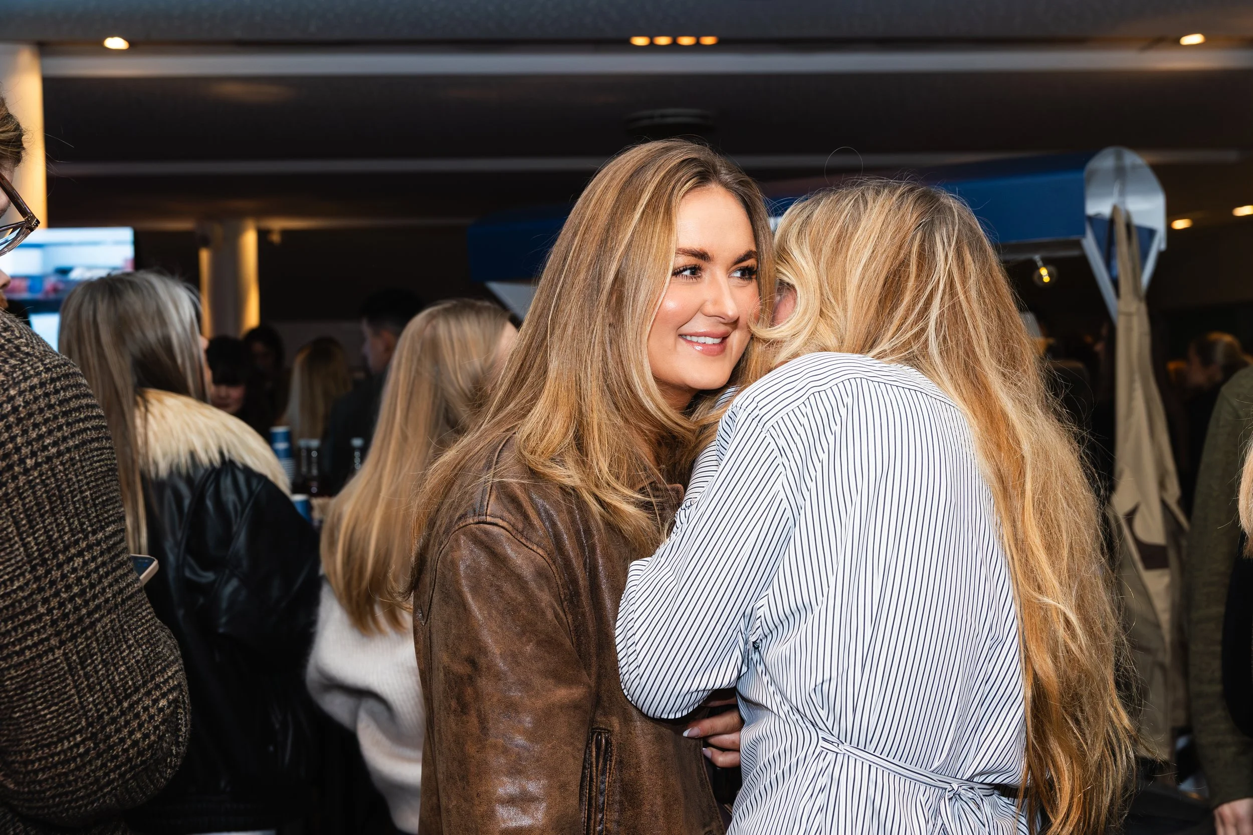 Two women with blonde hair embracing each other at a social gathering or event indoors, smiling and looking happy.