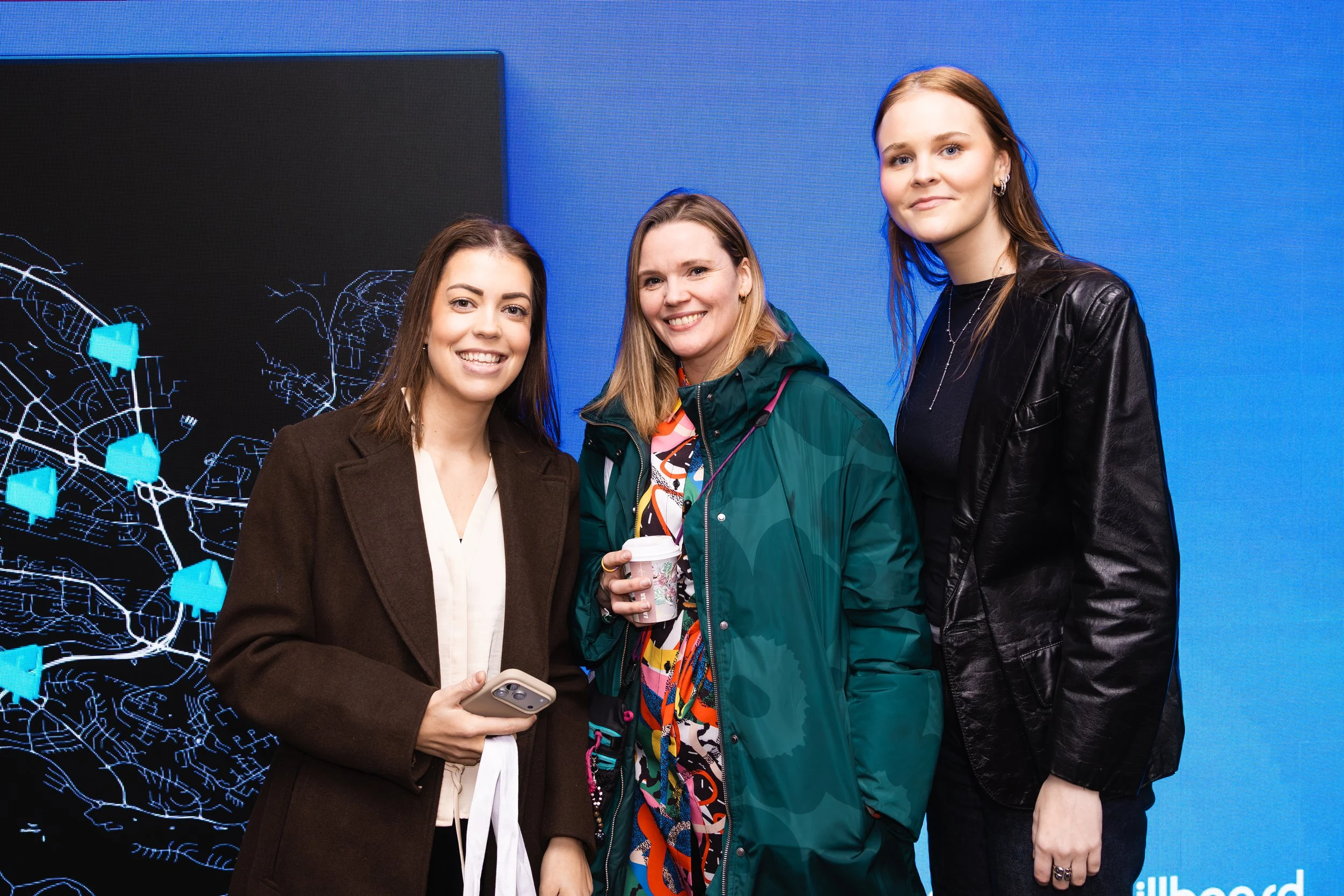Three women standing together indoors, smiling, with a large digital map or display in the background.