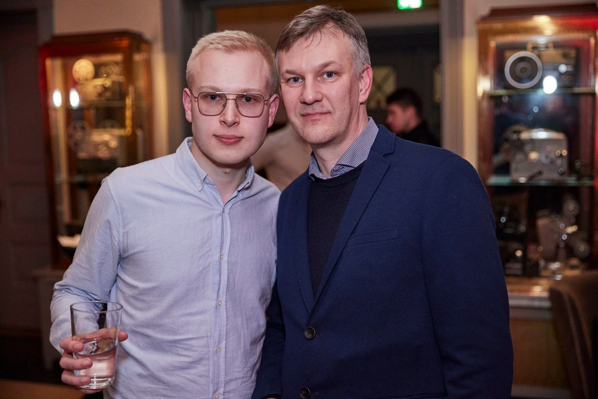 Two men standing close together at a social event, one holding a glass of clear liquid with ice, in an indoor setting with wooden display cases and blurred background.