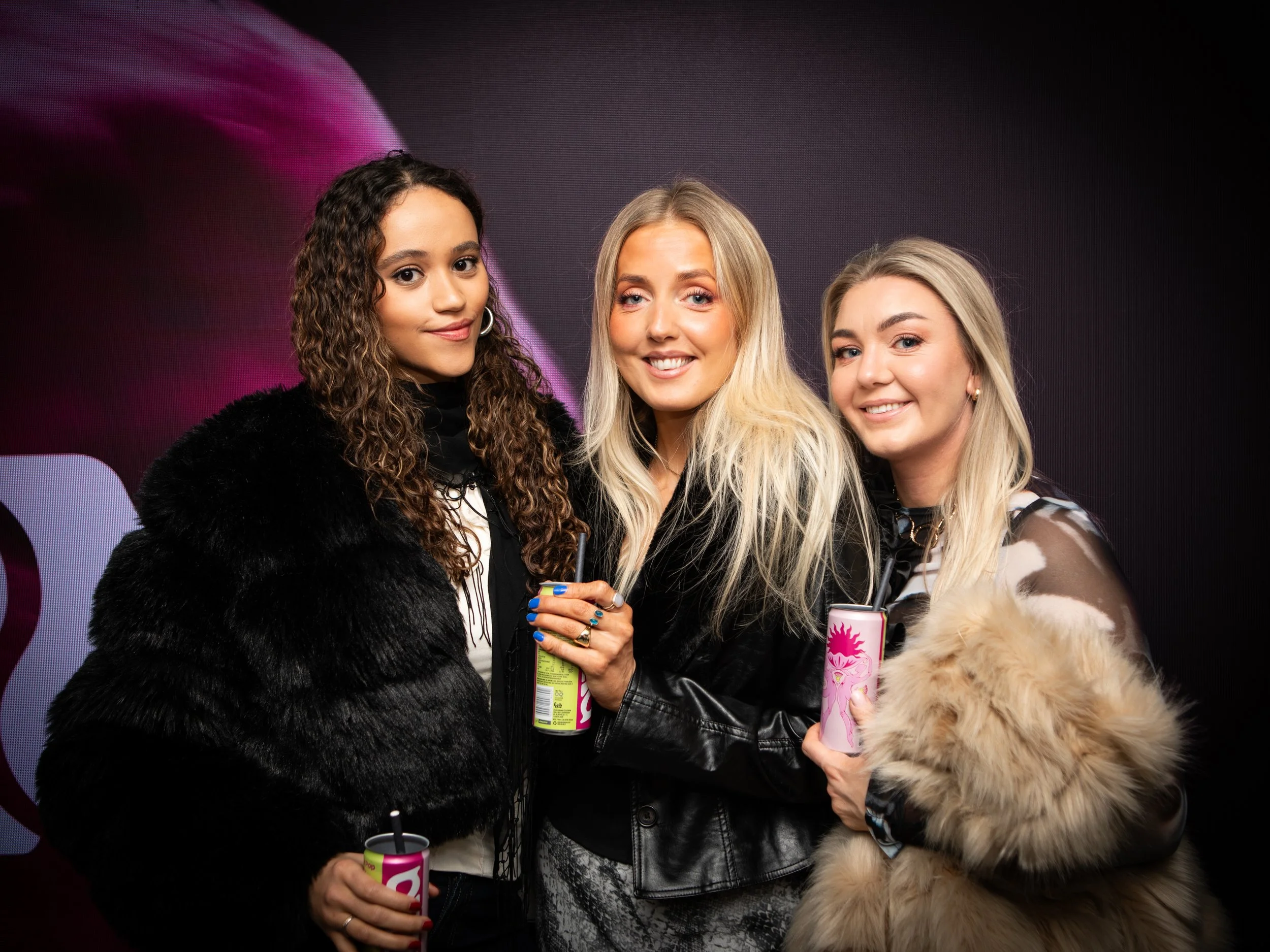 Three women standing together, smiling, holding pink cans with straws, dressed in stylish clothing with a dark background.