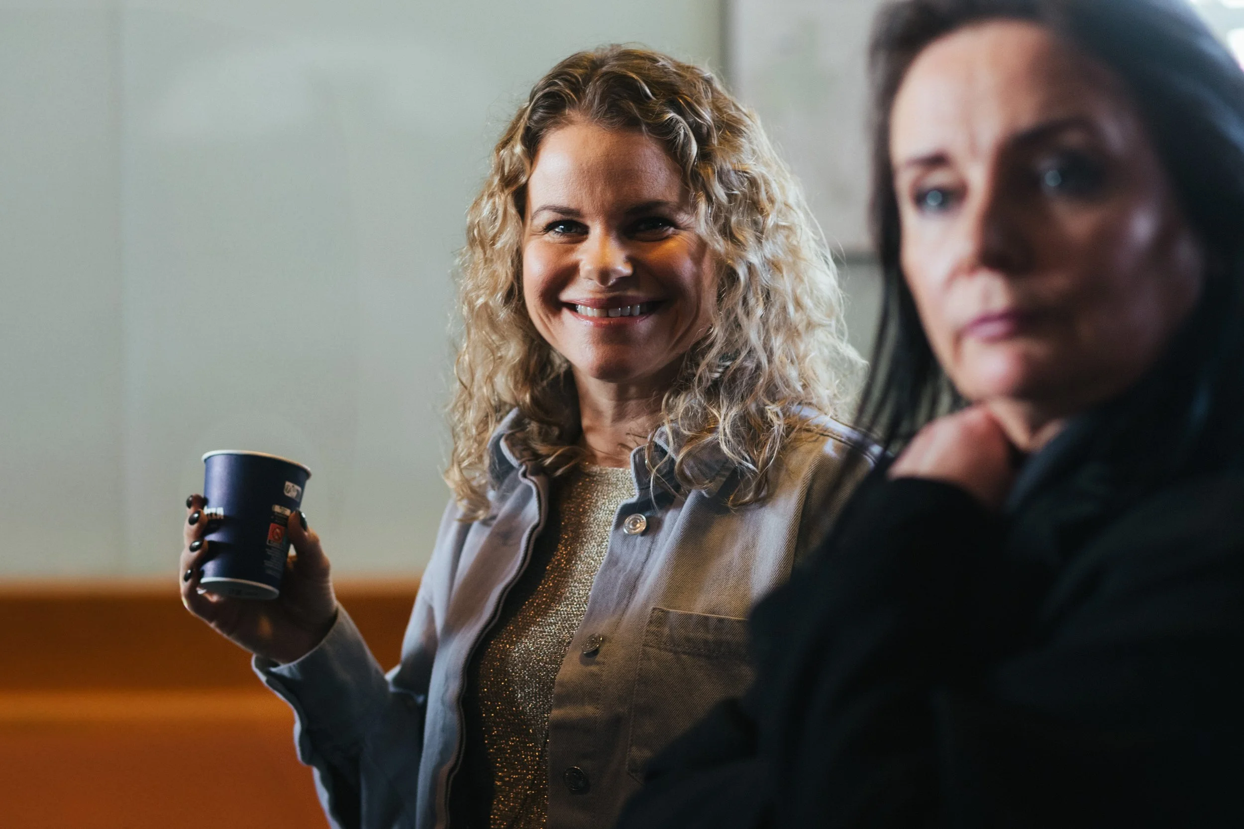 Two women sitting at a table, one smiling and holding a coffee cup, the other looking away.