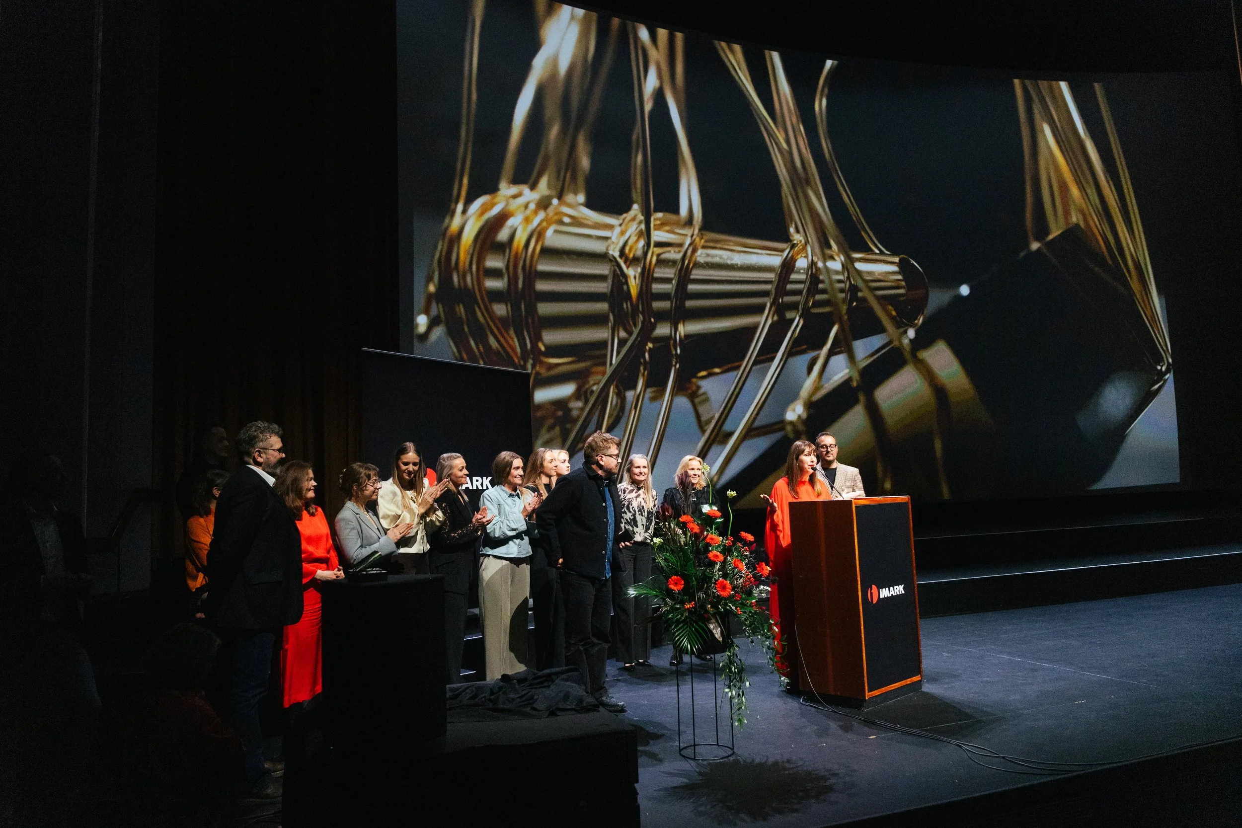 A group of people standing on a stage at an event, with a large screen behind them displaying an abstract gold and black image. There is a flower arrangement on a stand in front of a podium with a microphone.
