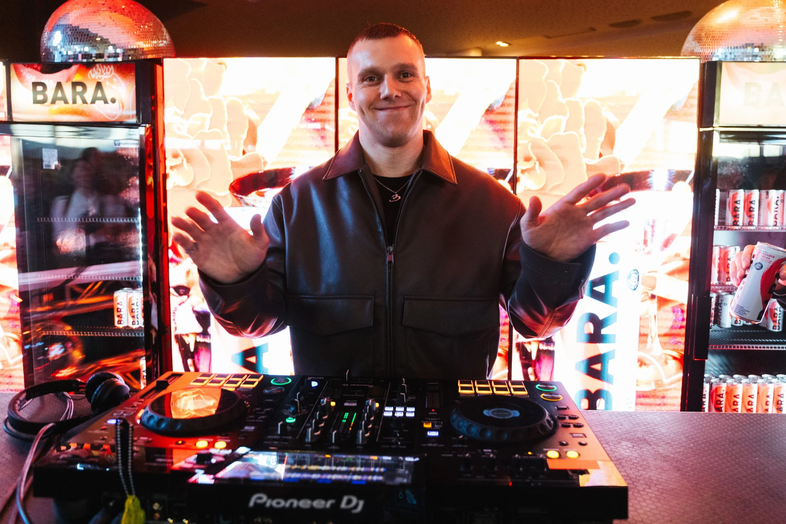 A man standing behind a DJ mixing console with a colorful, bright backdrop and illuminated display screens in the background.