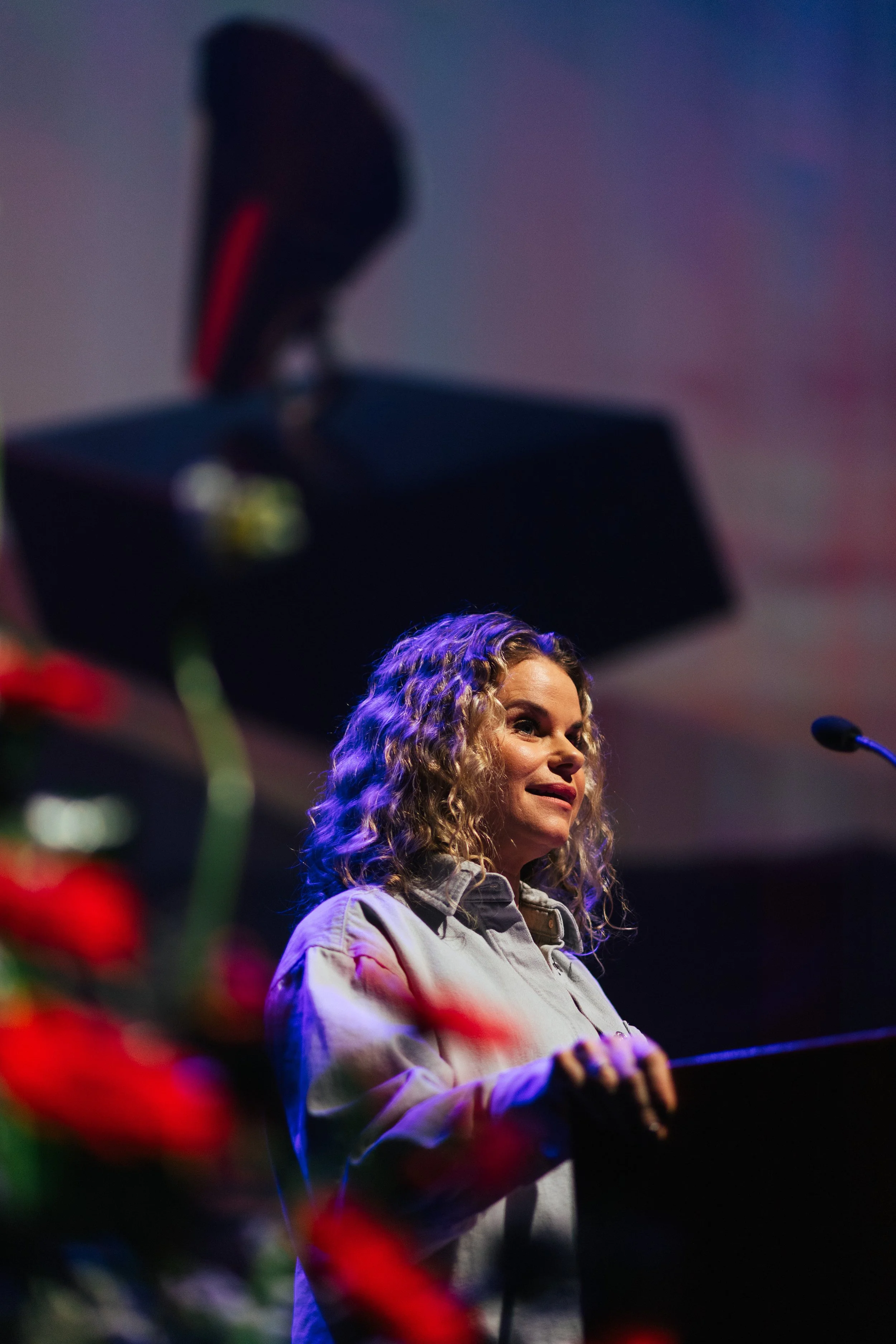 A woman with curly blonde hair standing at a microphone, wearing a light-colored jacket, with blurred red flowers in the foreground and a dark background.