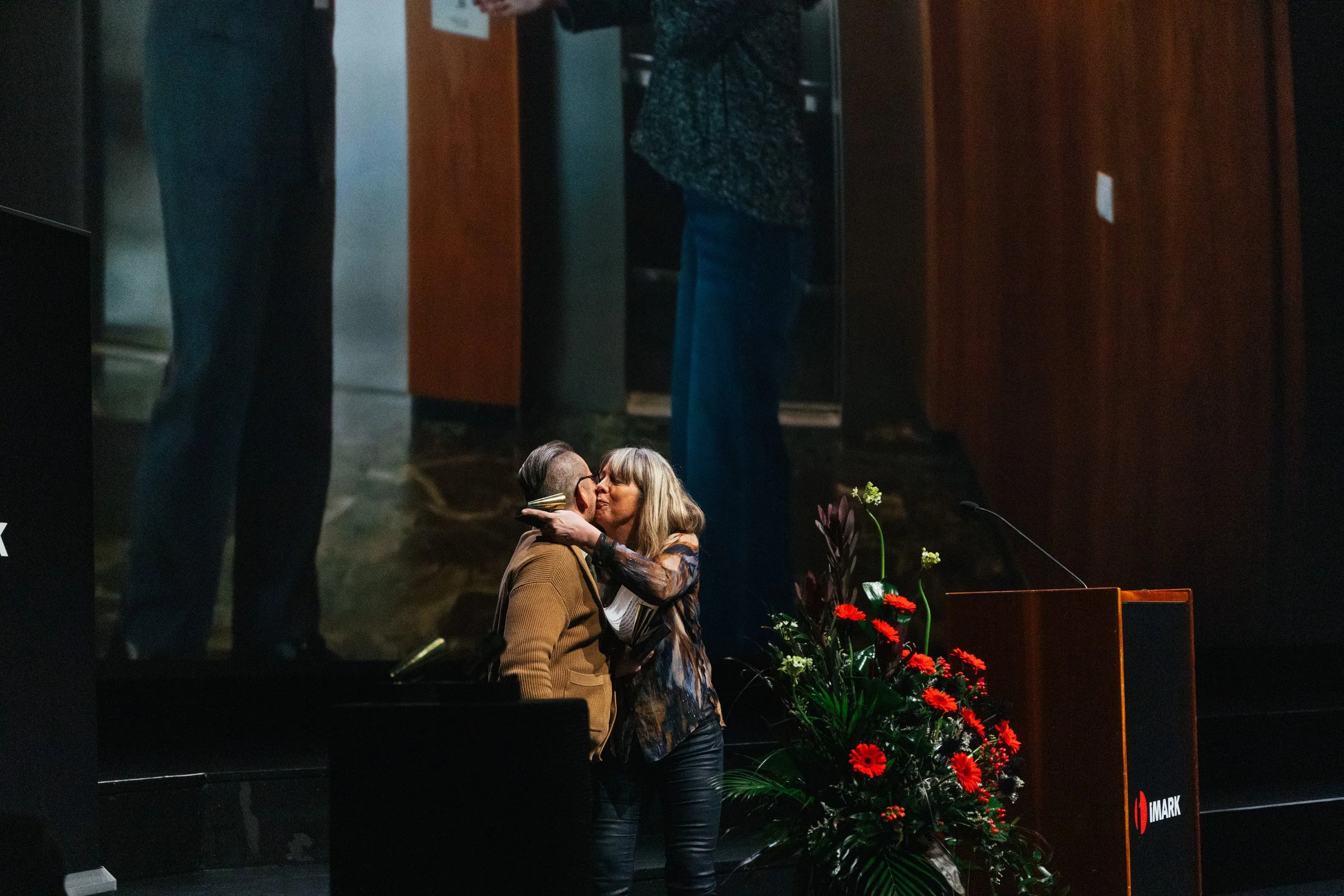 A woman and a man sharing a kiss, standing beside a woman in a floral dress, on a stage with a podium and red flowers, possibly during a formal event.