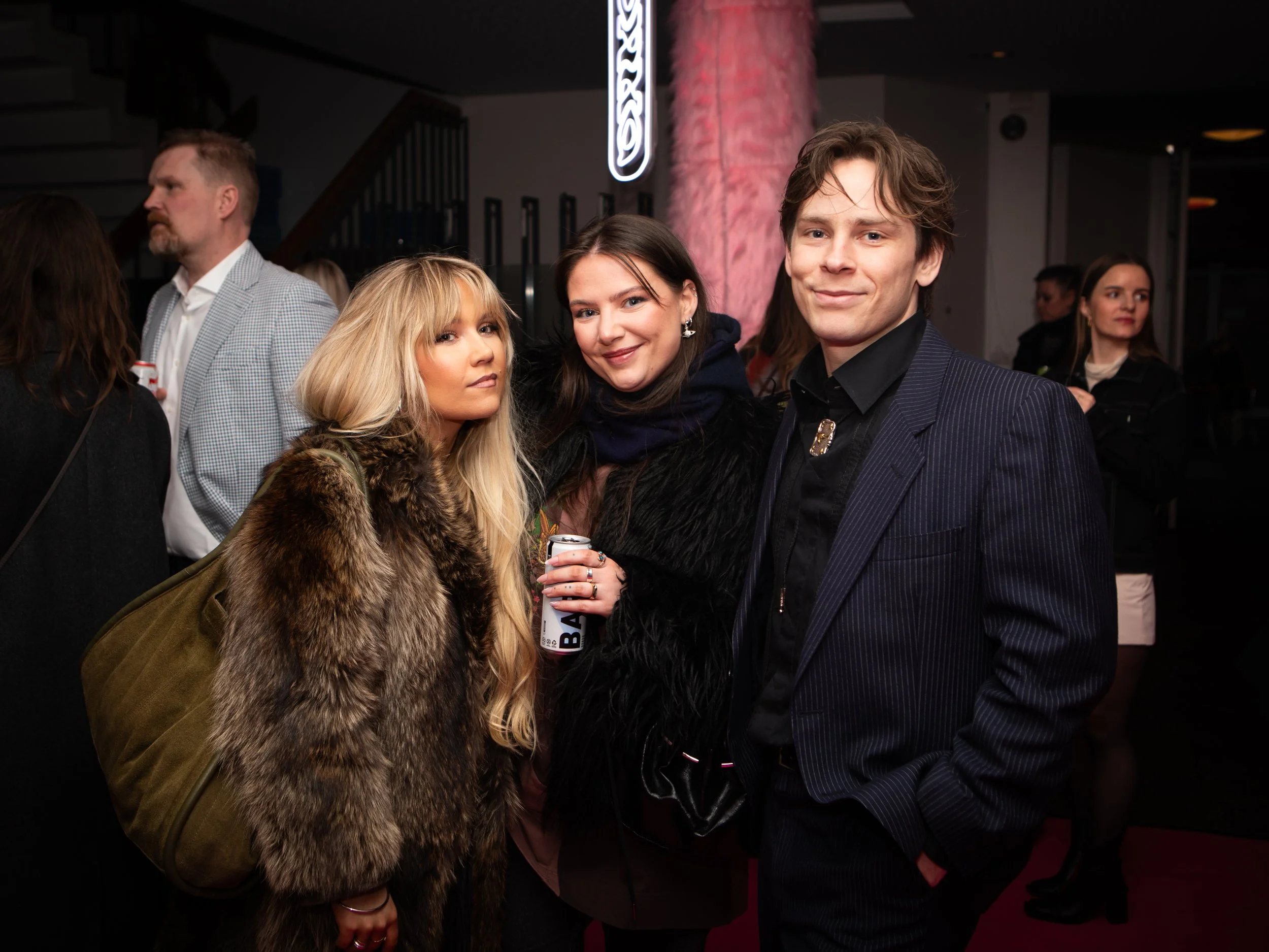 Three people posing for a photo at a social gathering with other people in the background.