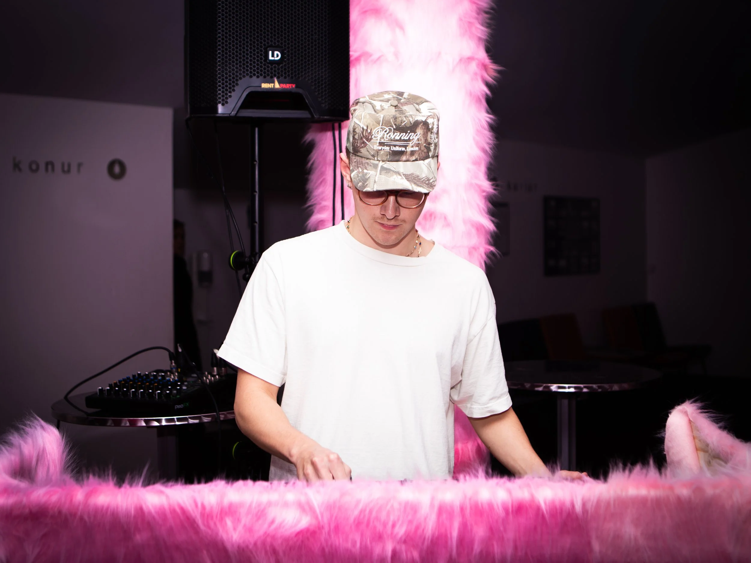 A young man wearing glasses and a camouflage cap that says "Ronnings" stands behind a table with pink fluffy fabric, in a room with a DJ mixer and a speaker in the background.