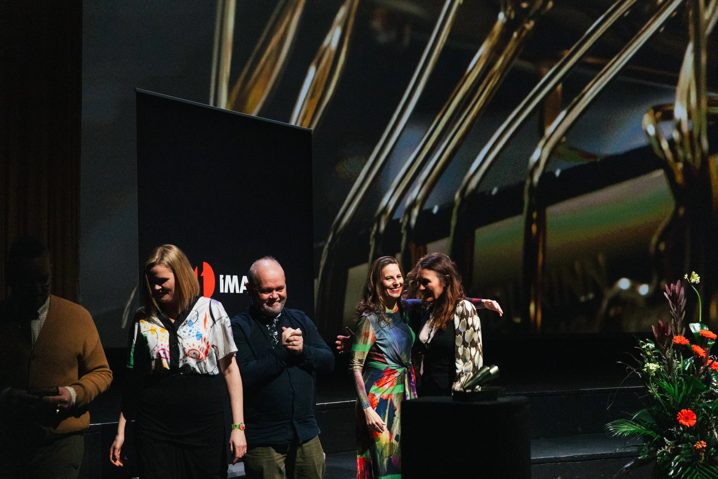 Group of five people standing on stage at an event, smiling, with flowers in front. A black banner with red and white text is in the background. The setting appears to be a formal or professional gathering.
