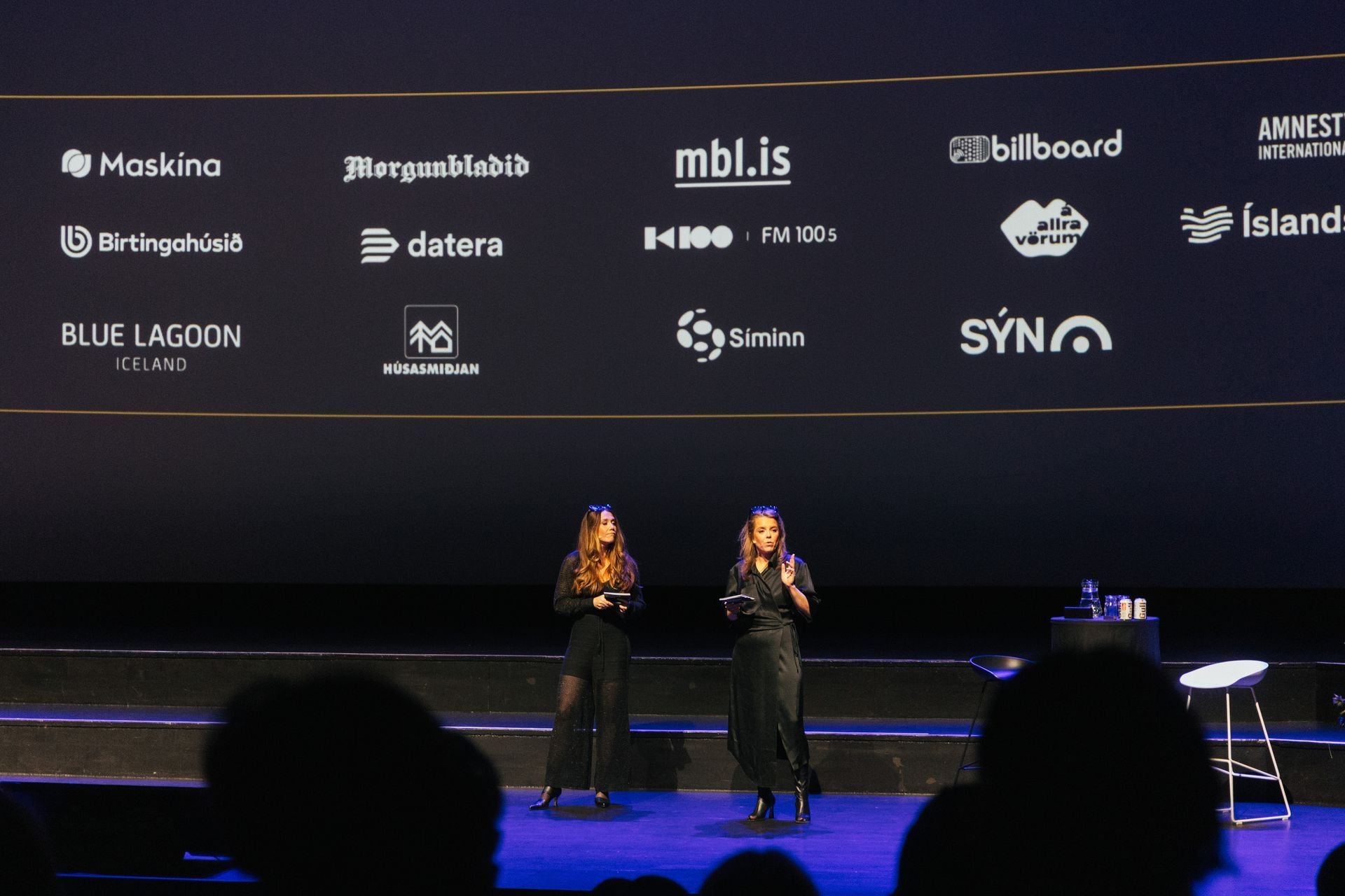A stage with two women, one speaking and the other possibly holding a microphone, with a large screen behind them displaying various logos and website names.