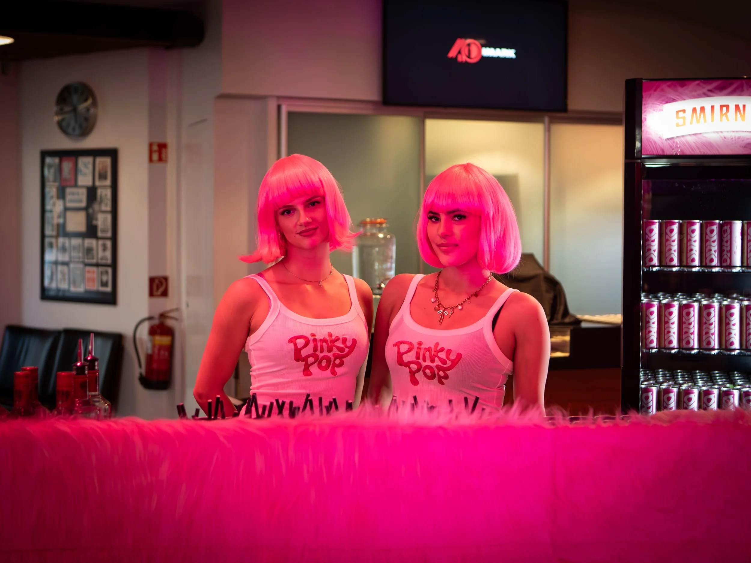 Two women with short pink hair and matching pink tank tops that say 'Pinky Pop' stand behind a pink furry counter in an indoor space, with soda cans and bottles on display and a mural in the background.