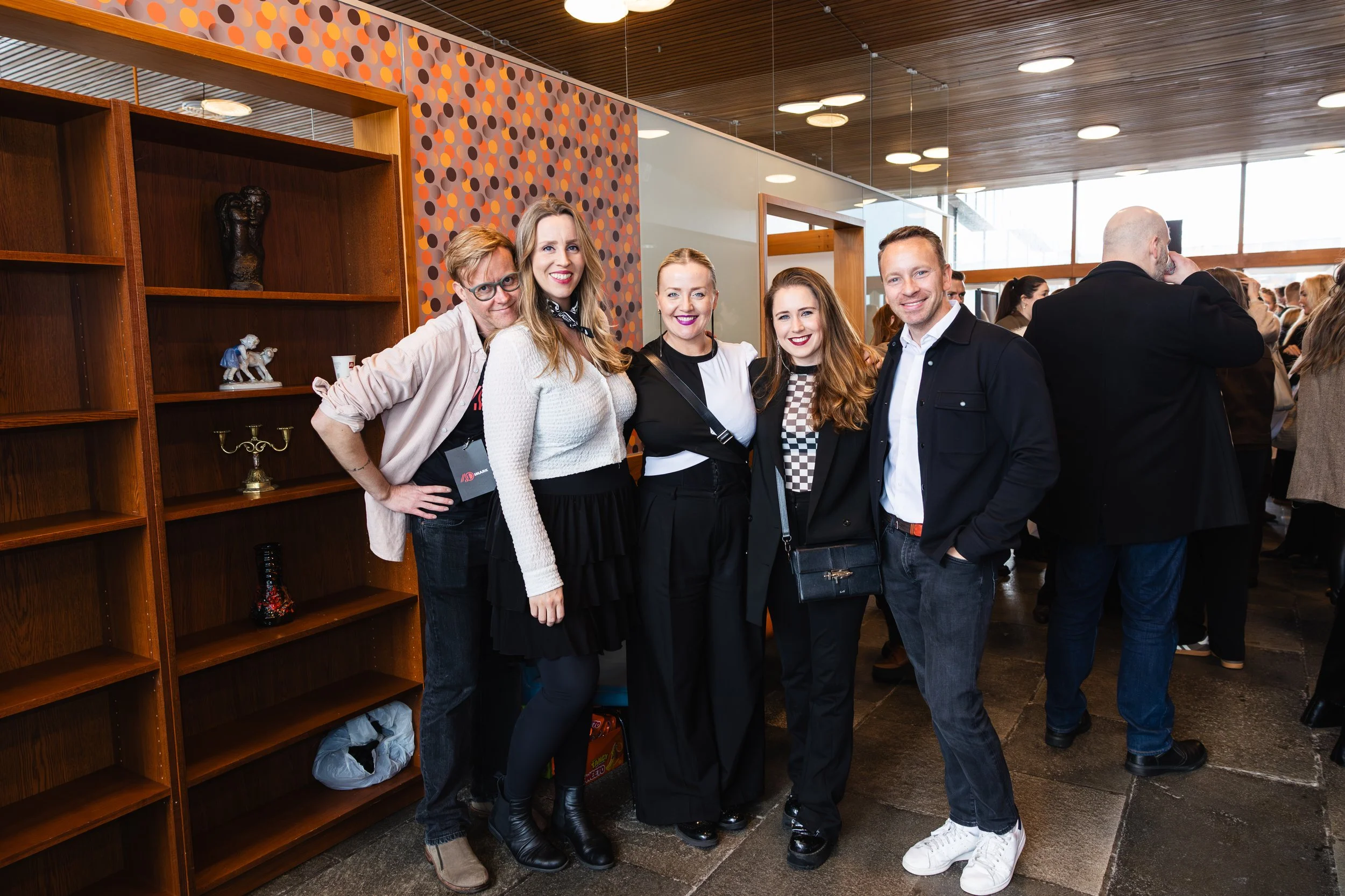 A group of five people standing together in an indoor setting, smiling at the camera with a wooden shelf and a decorative wall in the background.