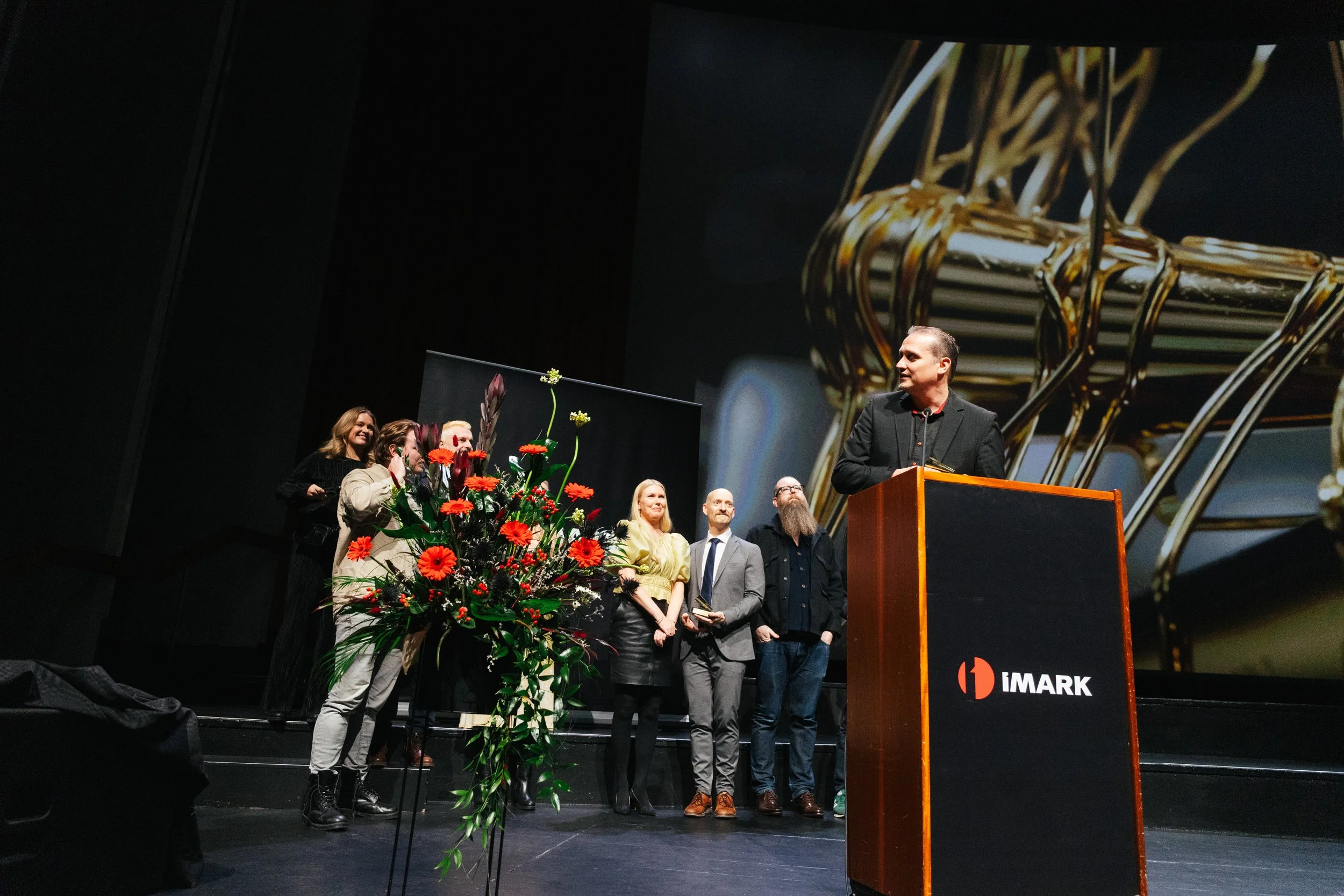 Group of people standing on a stage during an award ceremony, with a large floral arrangement in front, a podium, and a presenter speaking to the audience, with a large screen showing a close-up of a gold trophy in the background.