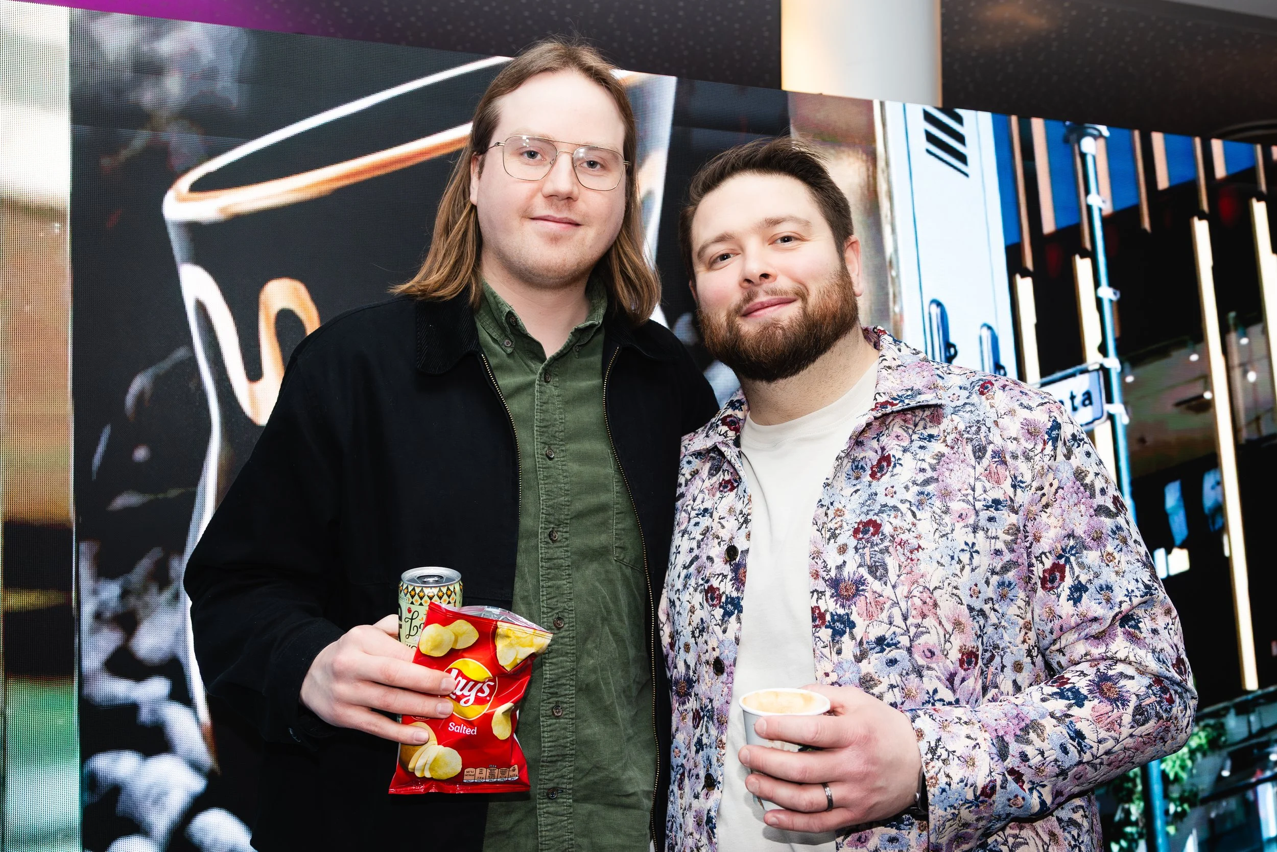 Two men standing together at an event, one holding a bag of Lay's salted potato chips and the other holding a cup of drink, with a large digital display in the background.