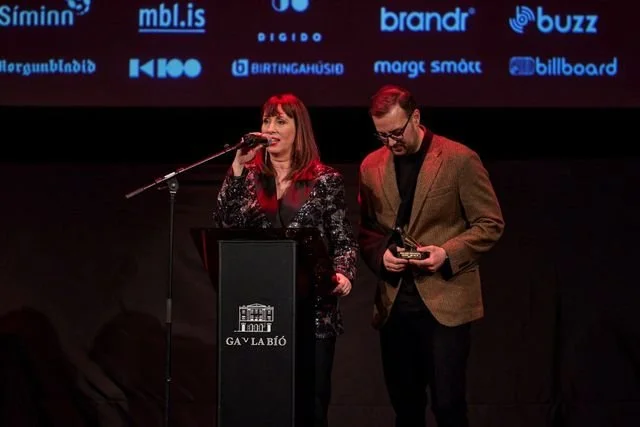 A woman and a man standing on a stage at an awards event, with the woman speaking into a microphone at a podium. The backdrop displays logos of various sponsors.
