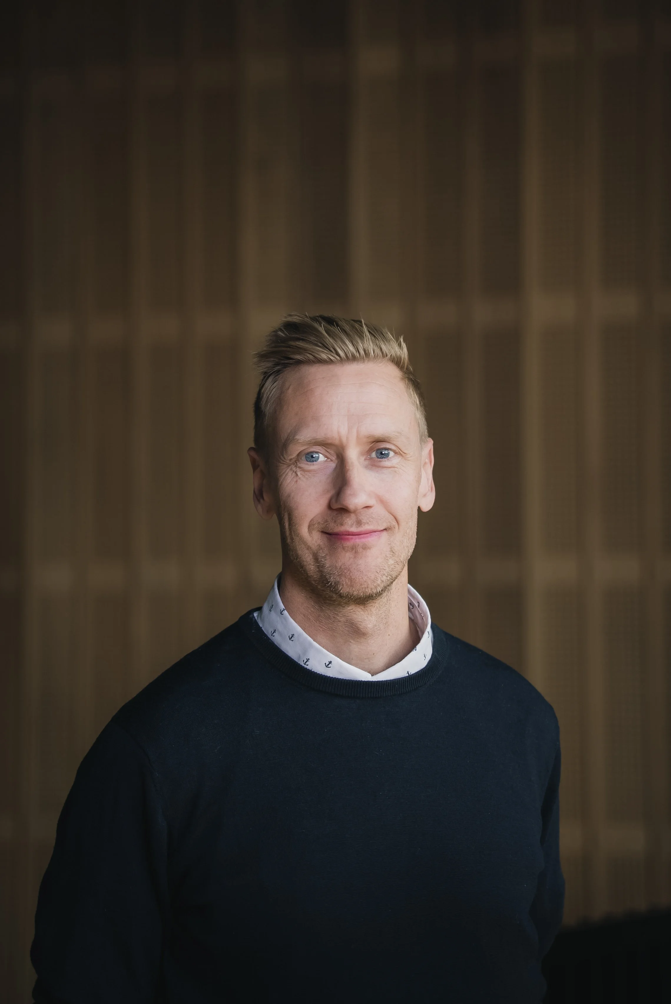A portrait of a man with short, styled blonde hair, blue eyes, and light skin, wearing a dark sweater over a collared shirt with anchor patterns, standing against a blurred background with vertical wooden panels.