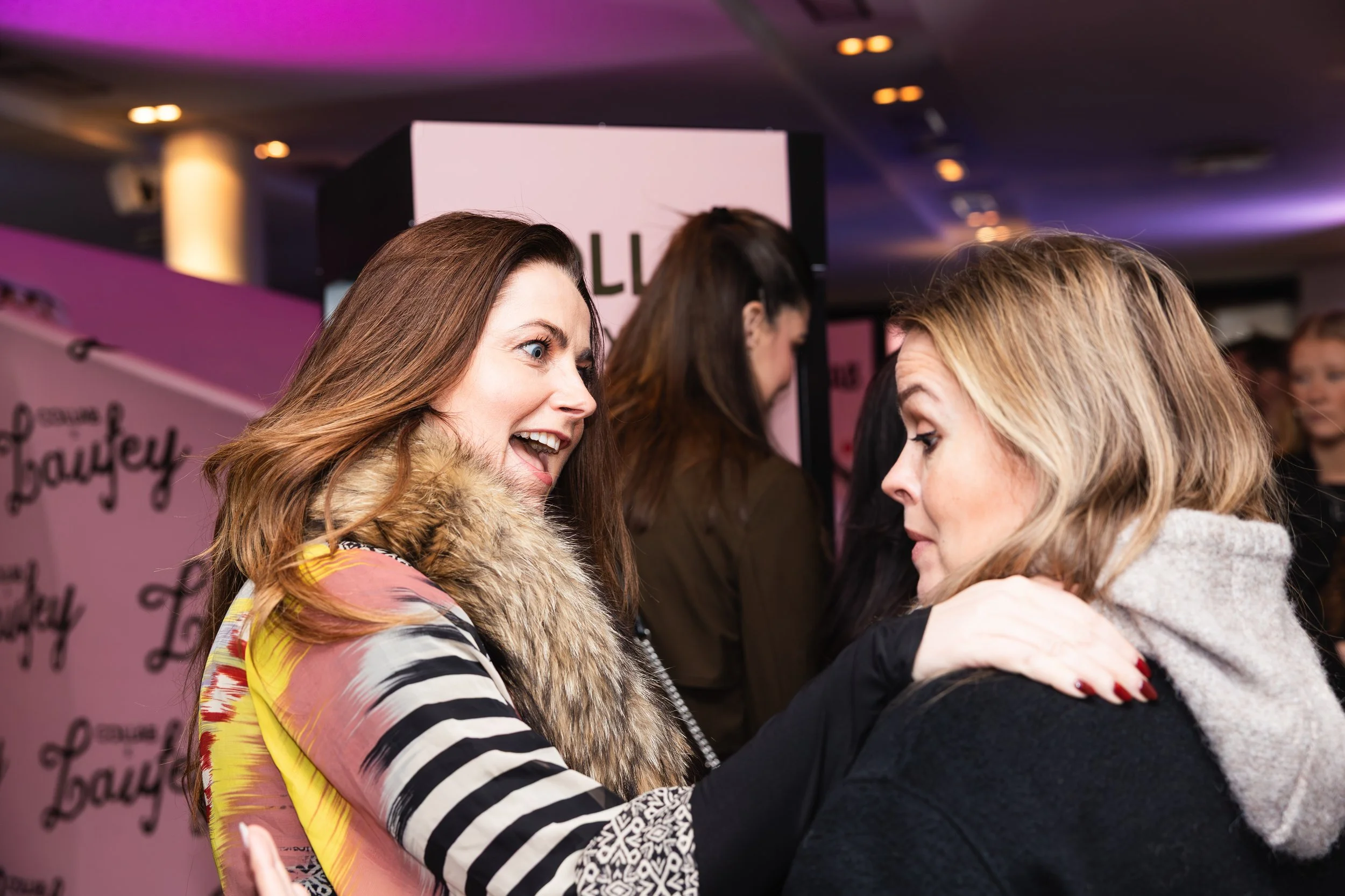 Two women hugging and talking at an event, with a pink background and other people in the background.