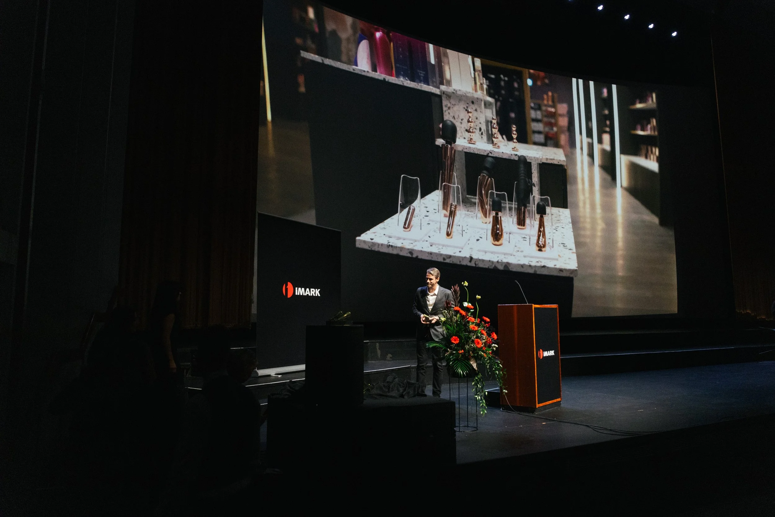 A person standing on a stage at a presentation with a large screen behind them displaying a close-up image of amber glass bottles and spray bottles. The stage is decorated with a floral arrangement, and the presentation is associated with the IMARK b