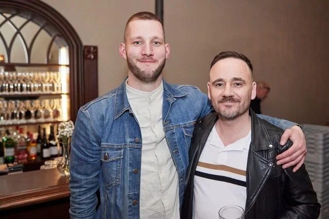 Two men posing together at a social gathering, one with a beard wearing a denim jacket, and the other with facial hair wearing a leather jacket, in a room with a bar and decorative mirror in the background.