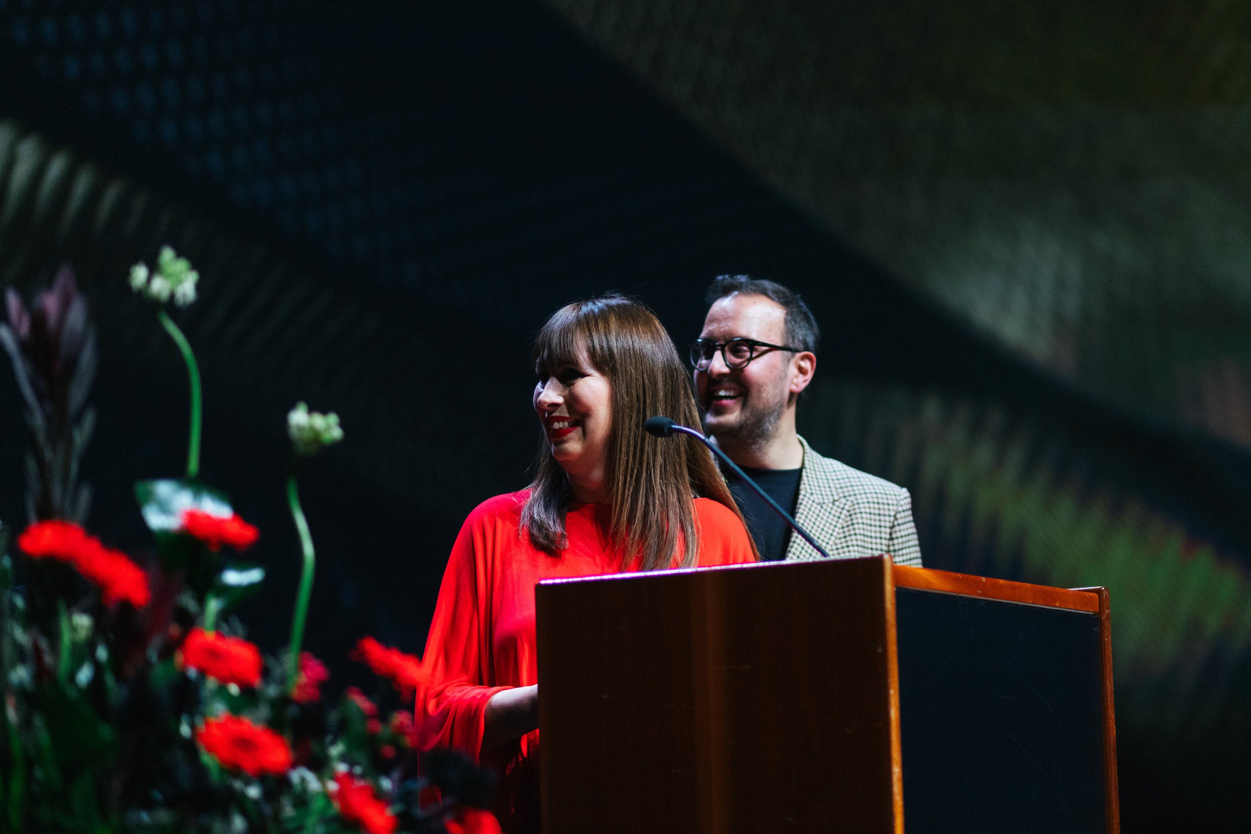 A woman with long brown hair and red lipstick wearing a red dress is standing at a wooden podium, smiling and speaking into a microphone, with a man wearing glasses and a checkered blazer standing behind her, smiling. There are flowers in the foregro