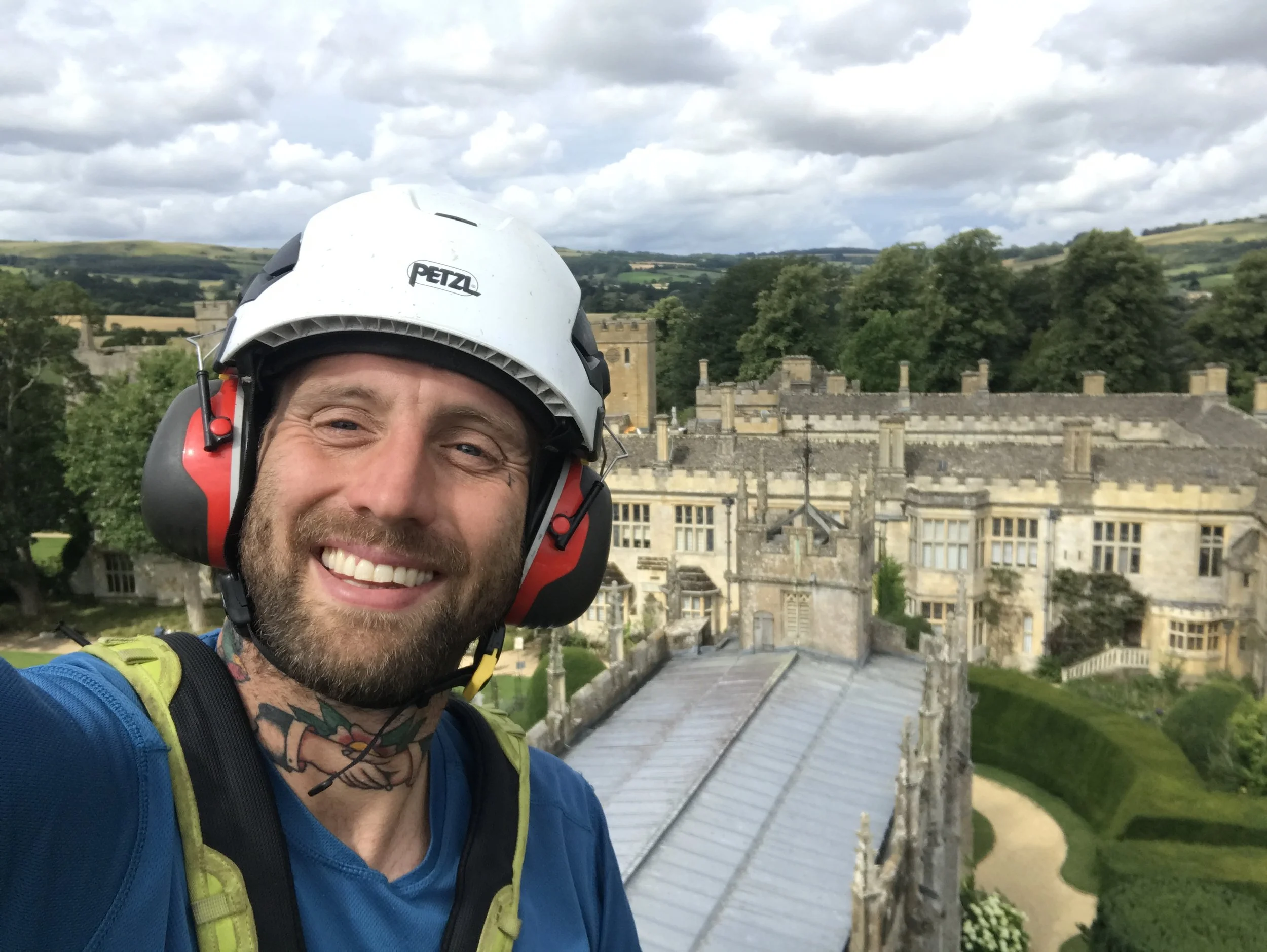 Smiling man wearing helmet and backpack taking a selfie on a rooftop with a castle and rolling green hills in the background.