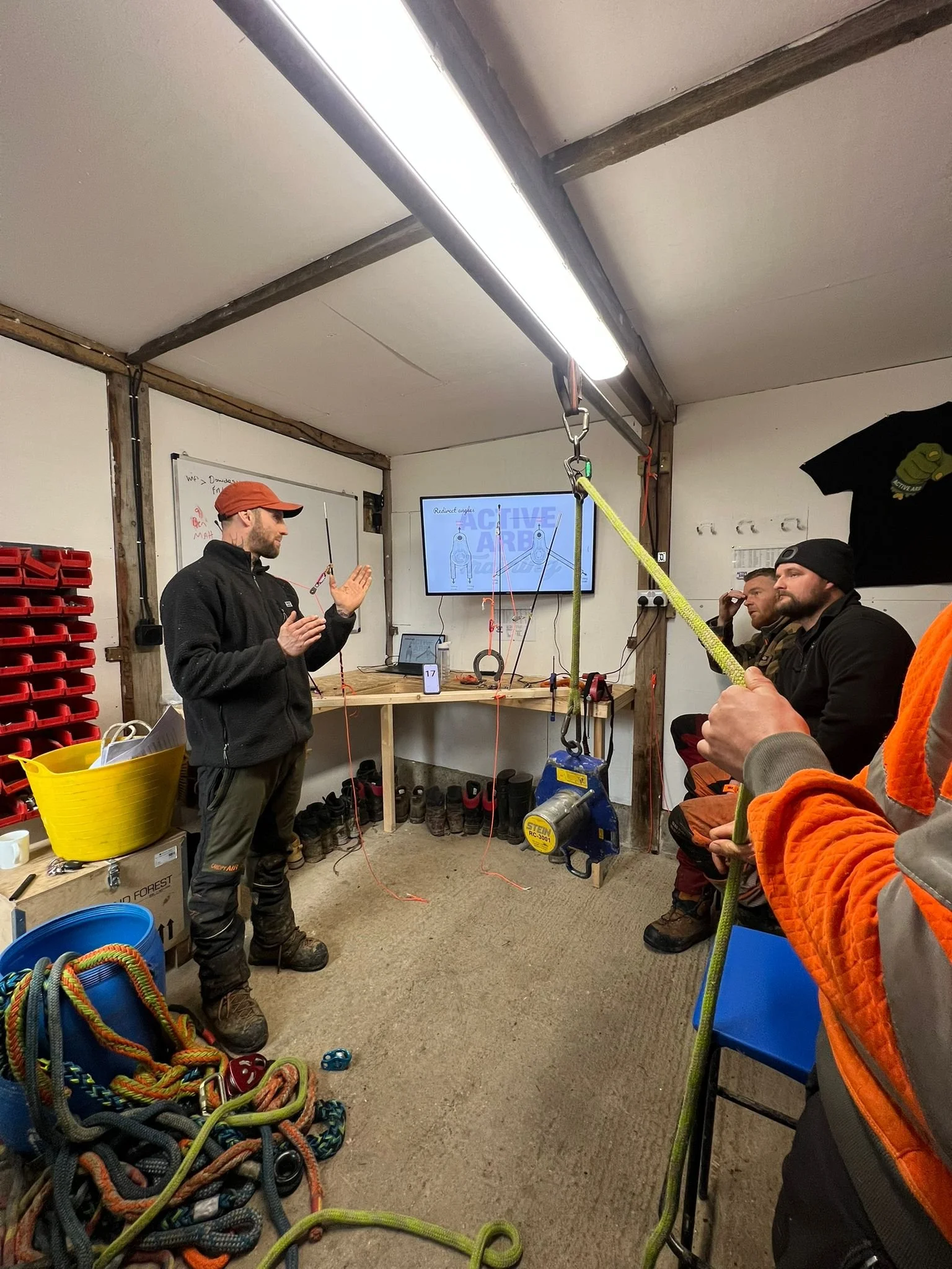 A group of people inside a workshop or training room, listening to a man in a black jacket and red cap giving a presentation on climbing or rope rescue, with ropes and climbing gear in the foreground.
