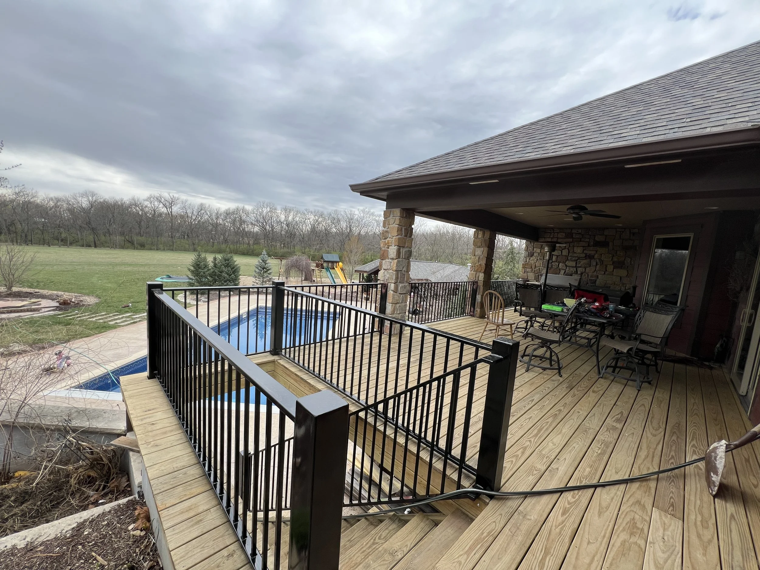 A backyard patio area with a wooden deck, black metal railing, and a covered porch with outdoor furniture. There is a swimming pool in the yard, a children's playground, and a large open grassy area with trees and overcast sky.