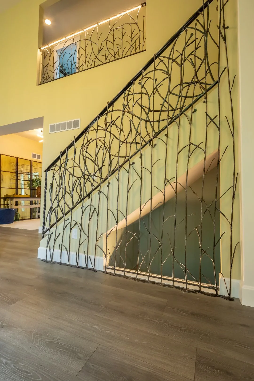 Interior view of a staircase with an artistic black metal railing resembling tree branches, against a light green wall. The staircase is illuminated by LED strip lighting, and the floor is wooden with a natural finish.